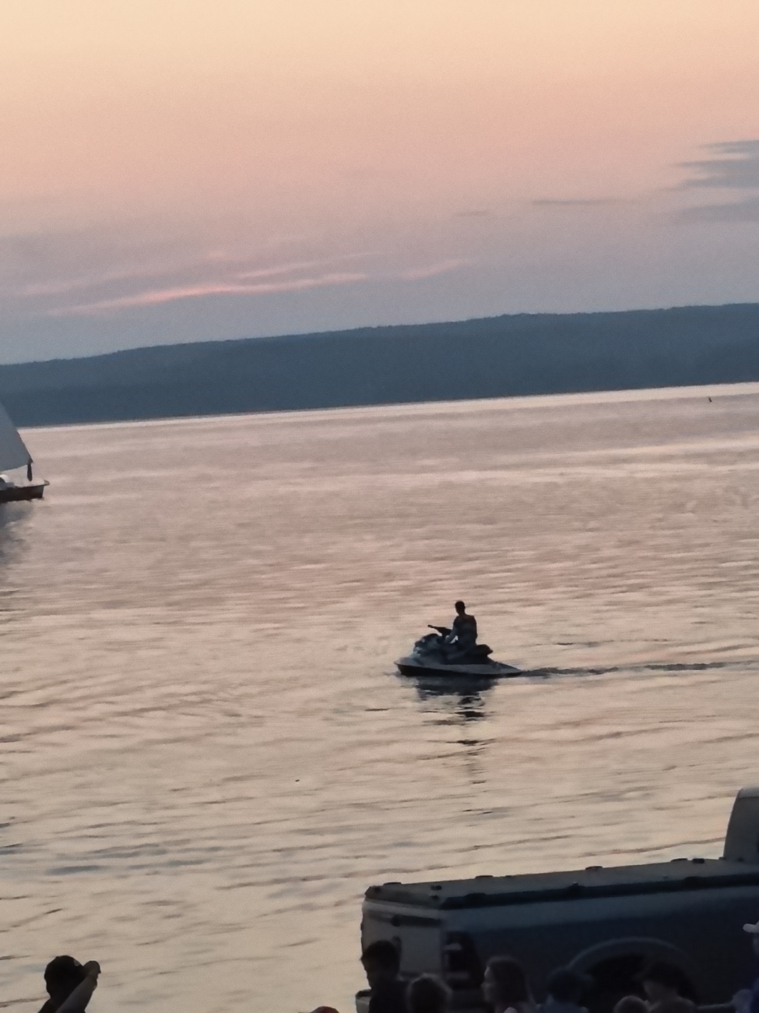 The image depicts a serene twilight scene on a body of water, possibly Lake Osa in Russia. A lone individual is riding a jet ski across the calm water, leaving a subtle wake. In the distance, a sailboat is visible, its white sail stark against the muted pastel hues of the sky. The horizon is defined by a dark, tree-lined landmass. In the foreground, a portion of a pickup truck bed is visible, suggesting the vantage point is from the shore or a nearby vessel. Silhouettes of people are present at the bottom of the frame, indicating onlookers or participants in an event. The overall atmosphere is peaceful and tranquil, with the soft light of dusk casting a warm glow over the water and sky.