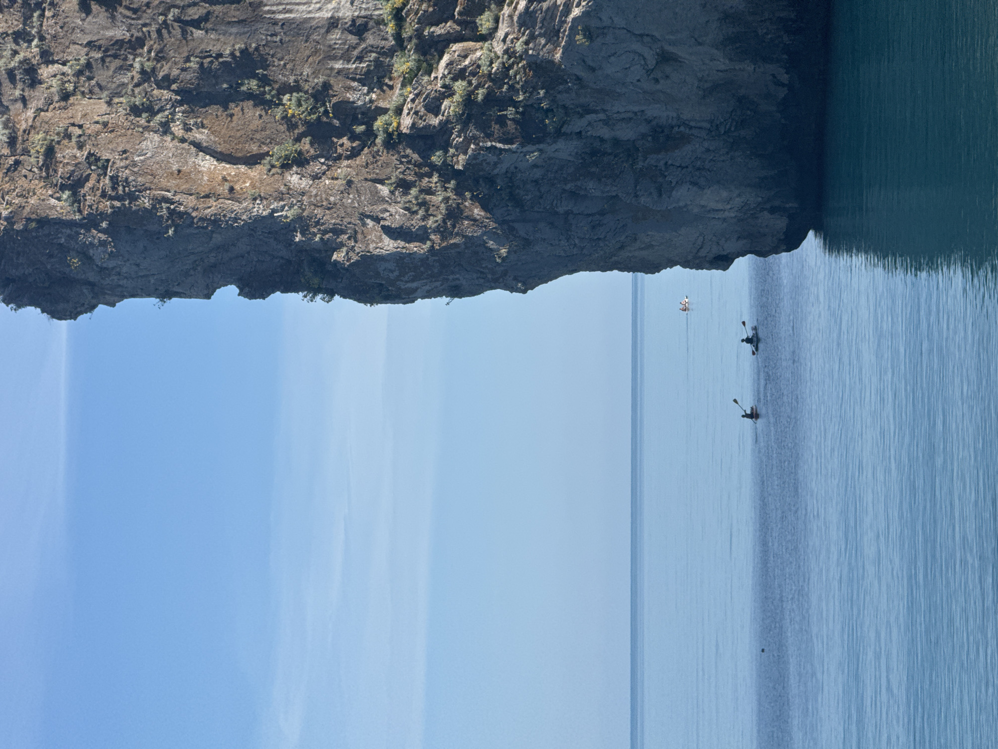 The image depicts a scenic view of a rocky cliff face overlooking a calm body of water. The cliff is rugged and covered with sparse vegetation. The water is a deep blue, with subtle ripples indicating a tranquil atmosphere. Three individuals are visible in the water, each in a small boat, appearing to be kayaking or paddling. They are spaced out, suggesting a leisurely activity.
The sky is a clear, bright blue, suggesting fair weather, likely during the daytime. The light appears bright, casting subtle shadows and highlighting the textures of the rock. The composition of the image, with the imposing cliff and the expansive water, creates a sense of natural beauty and serenity. The presence of the kayakers adds a human element to the otherwise natural landscape, emphasizing the opportunity for recreation in this environment. No visible text is present in the image.