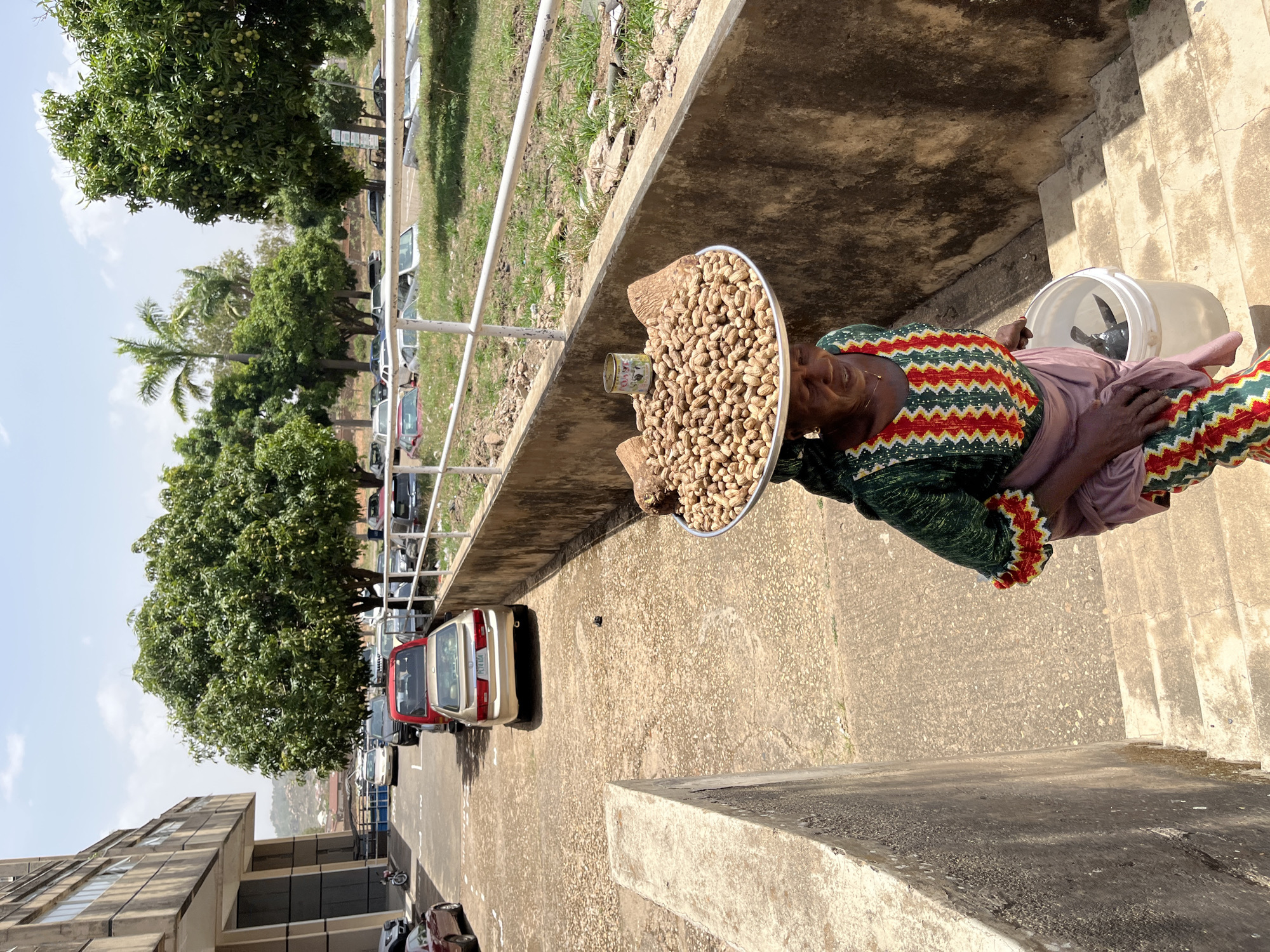 An elderly woman is selling peanuts in Jos, Nigeria. She sits on steps next to a building, holding a large tray piled high with peanuts and a tin can. Beside her is a white bucket containing something wrapped in a black plastic bag. She is wearing a green and red patterned top, a lavender skirt, and patterned pants. The background includes a street with parked cars, trees, a building, and a partial view of a hillside. The setting seems to be in the vicinity of a university or public building, given the architecture and parking area.