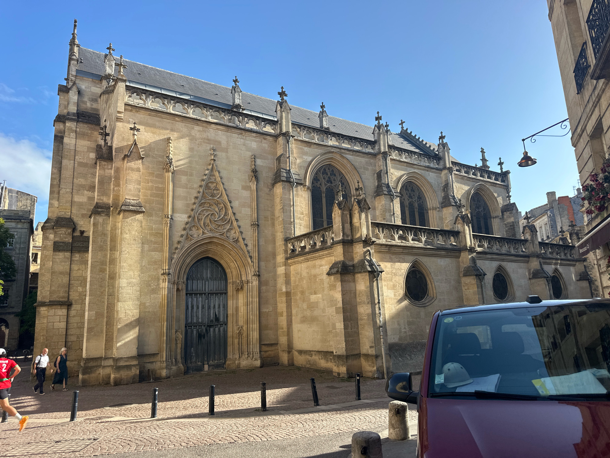 The image shows the exterior of a large, historic stone church with a tall, pointed roof in Bordeaux, France. The church features ornate architectural details, including arched windows with gothic tracery and decorative stonework around the entrance. The church is situated on a cobblestone street, and to the right, a portion of a modern maroon van is visible in the foreground, partially obscuring the view of a building. Two people are walking on the sidewalk in front of the church, and a person wearing a red shirt with a number is running in the foreground. The sky is a clear, bright blue with some scattered clouds. The sun casts strong shadows, indicating a sunny day. The architecture suggests the church is a significant historical landmark in the city.