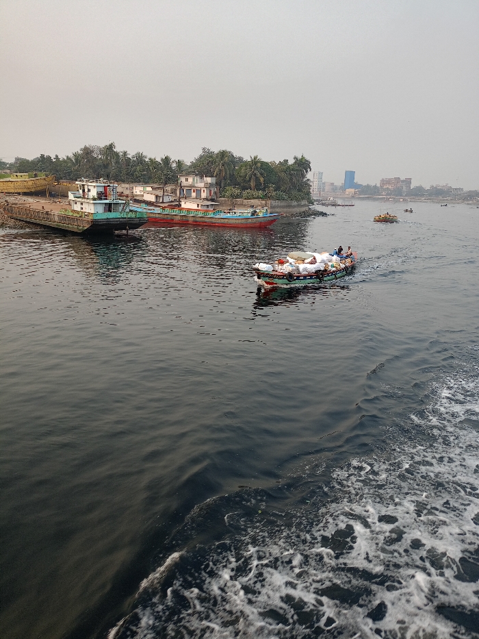 The image captures a bustling river scene in what appears to be Zazira, Bangladesh, under an overcast or hazy sky. The central focus is a wide waterway with dark, rippling water.
In the mid-ground, a motorized longboat, painted green and red, moves from right to left, heavily laden with numerous white sacks and other cargo. Two or three individuals are visible on board, likely crew or passengers. The boat leaves a distinct white, foamy wake behind it. In the far distance on the water, several smaller boats are indistinctly visible, suggesting further river traffic.
On the left bank, a collection of structures and vessels is visible. A large, flat-bottomed cargo boat or barge, painted light blue and white, is either docked or grounded near the shore. Behind it, a longer, narrower boat with a red hull and blue top is also moored. Further left, what looks like a skeletal yellow boat frame or hull is visible on the bank. The riverbank itself is lined with lush green trees, including palm trees, and residential-style buildings with light-colored walls and dark roofs are nestled among the foliage.
The background shows a distant cityscape with a cluster of buildings, some taller structures, indicating an urban area across the river. The sky is a uniform light grey, suggesting either an overcast day or atmospheric haze, which diffuses the light and creates a soft, even illumination. The time of day appears to be daylight, likely morning or afternoon. No discernible text is visible on any of the boats or buildings. The scene depicts everyday life and commerce along a busy river.
