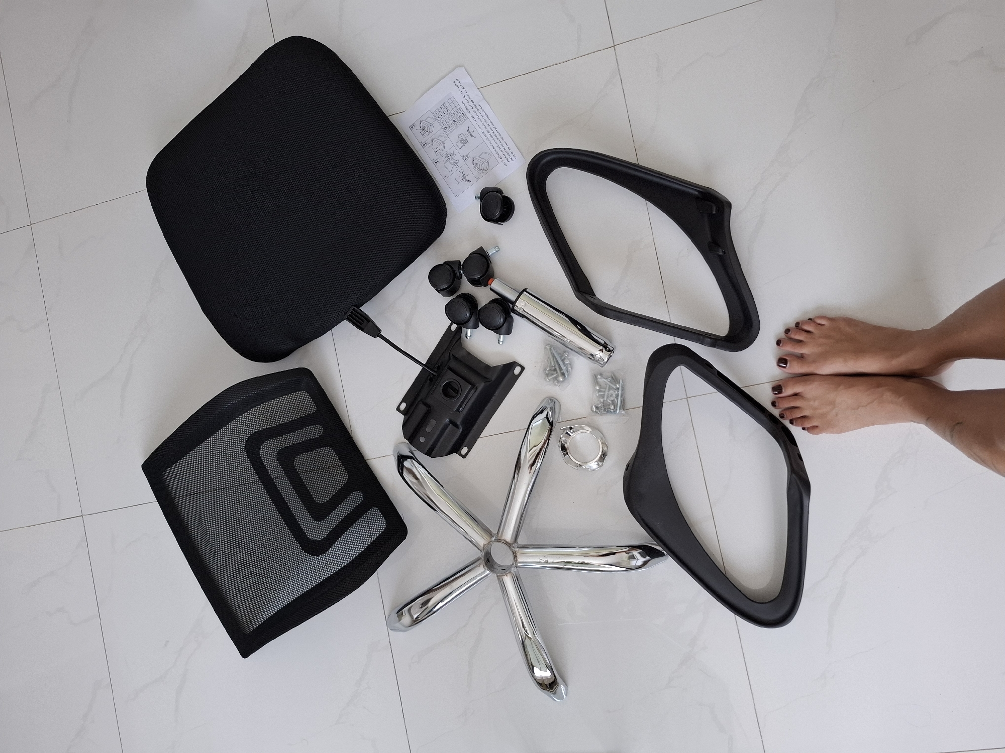 The image shows disassembled parts of an office chair laid out on a white tiled floor. The parts include a chrome five-star base, a gas lift cylinder, five caster wheels, a seat cushion, a mesh backrest, two armrests, a tilt mechanism, and a small instruction manual. A person's feet, with dark nail polish, are visible at the bottom of the frame, indicating a human presence. The lighting suggests an indoor environment, likely a room or hallway. The scene is static, with no active assembly or interaction occurring. The location is specified as Nha Trang Ward, Vietnam.