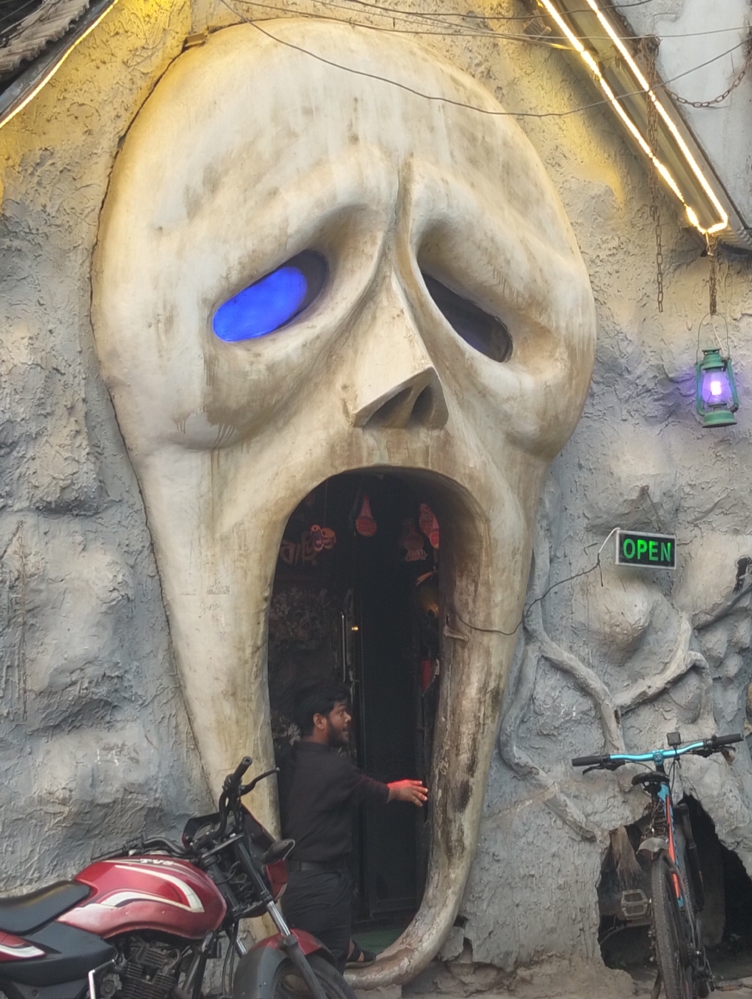 A large skull-shaped entrance, possibly to a haunted house or themed attraction, dominates the frame. The skull's eyes are illuminated with blue light, and its open mouth serves as the doorway. A man dressed in black stands in the doorway, his right hand extended outward. A red motorcycle is parked to the left of the entrance, and a bicycle is on the right. An "OPEN" sign hangs to the right of the entrance. A lantern hangs above the sign. The surrounding structure is designed to resemble rock or stone. This is in Dhaka, Bangladesh.