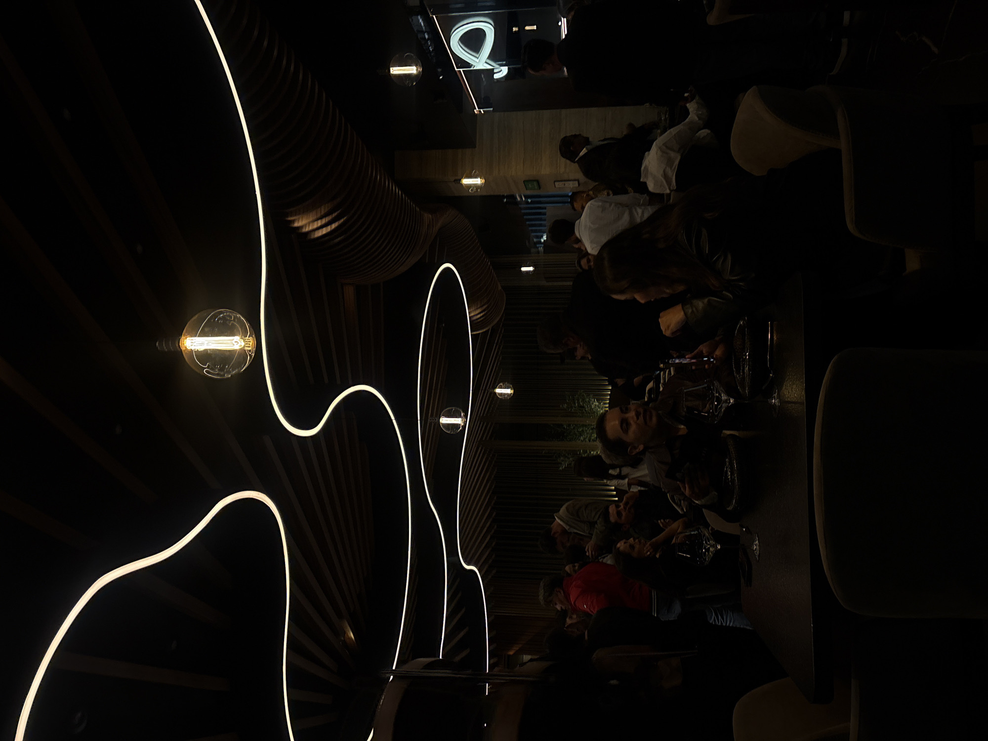 A dimly lit restaurant or bar scene shows patrons seated at tables, interacting with each other. The location is San Bernardino Tlaxcalancingo, Mexico. The interior design features a dark, possibly wooden, wall with linear, glowing white lights arranged in an abstract pattern. Round light bulbs hang from the ceiling. People are dressed in varied attire, including a red jacket and white shirts. They are likely eating and drinking, as suggested by the presence of glasses on the tables. The overall atmosphere appears to be social and relaxed.