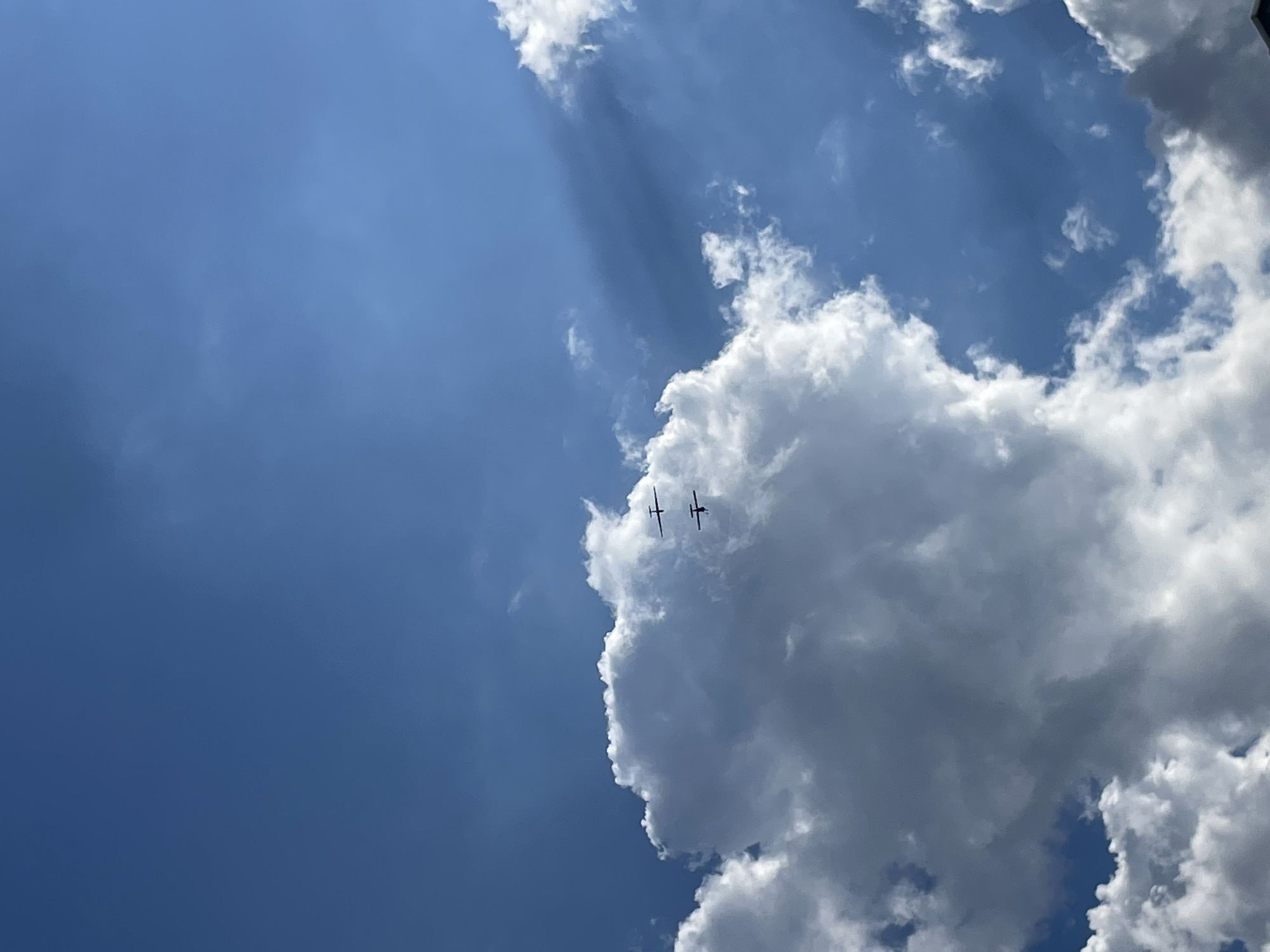 This media file captures a view of the sky on a partly cloudy day, likely during daylight hours. The sky is a vibrant blue, with several distinct clouds. One large, fluffy cloud dominates the right side of the frame, casting shadows and creating a sense of depth. There are also lighter, wispy clouds scattered across the sky, particularly in the upper left quadrant, where sunbeams are visible filtering through.
Two small, dark objects that appear to be airplanes are flying in formation near the large cloud. They are silhouetted against the bright sky, making their details difficult to discern. Their presence suggests an activity related to aviation, such as recreational flying or a demonstration.
The lighting indicates it is daytime, with the sun actively illuminating the clouds. The contrast between the bright sunlight and the shadows suggests a dynamic weather condition, moving from clear to cloudy. There are no discernible location cues or visible text within the image. The overall mood is one of openness and perhaps a touch of awe at the spectacle of flight against a dramatic sky.