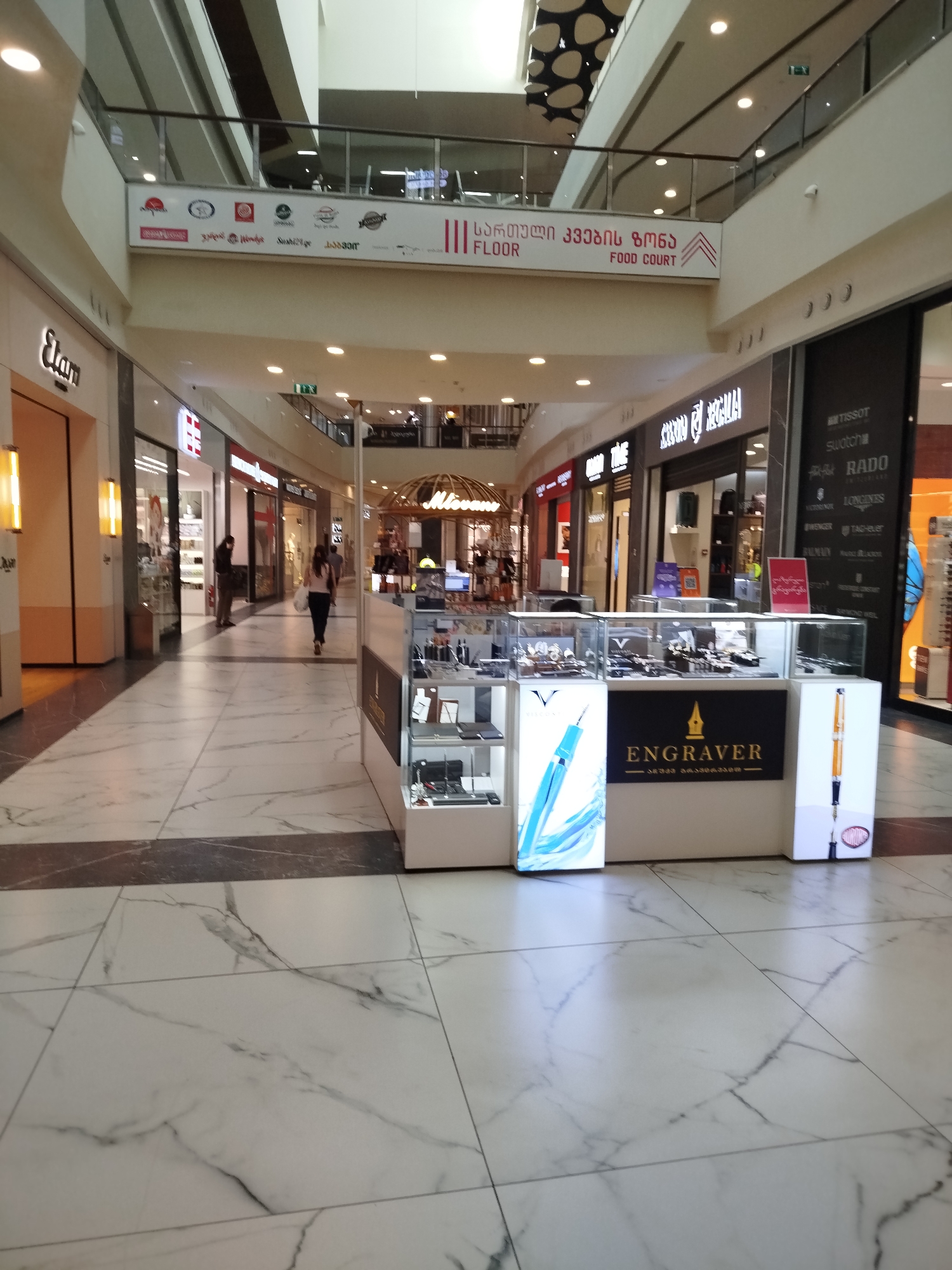 The media file shows an interior view of a modern shopping mall. The scene is brightly lit, suggesting it is daytime. The floor is made of large, polished marble tiles with intricate grey veining. In the foreground, there is a retail kiosk displaying various watches and jewelry in glass cases. The kiosk has prominent signage with the word "Engraver" in large, bold letters, along with Georgian script below. On either side of the main display are illuminated panels featuring images of elegant pens.
In the middle ground, several people are walking through the mall. One woman is walking away from the camera, carrying a white shopping bag, while another person stands near a shop entrance on the left. The mall architecture features multiple levels with balconies and railings, indicating a large, multi-story establishment. Shops line both sides of the walkway, with visible signage for brands like "Elan" on the left and a collection of watch brands such as Tissot, Swatch, Rado, and Longines on the right.
In the background, the mall continues with more shops and escalators. A sign above one of the walkways reads in Georgian script, "საქართველოს კვების ზონა" (Sakartvelos kvebis zona), which translates to "Georgia Food Zone," and also "FOOD COURT." This indicates the presence of a dining area within the mall. The overall atmosphere appears to be calm and orderly, typical of a shopping mall environment.