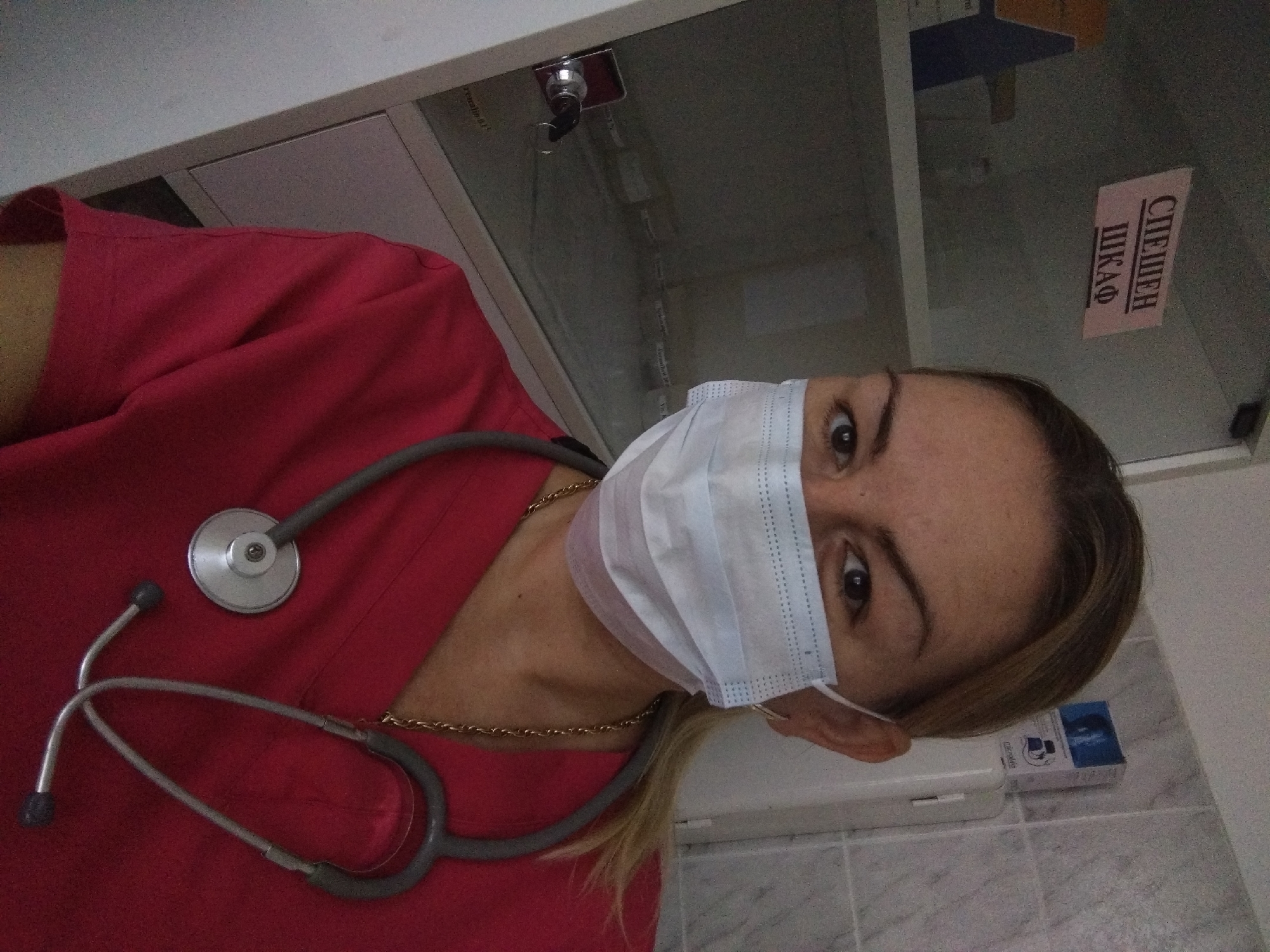 A woman wearing a red shirt and a light blue disposable face mask has a stethoscope around her neck. She is in what appears to be a medical or clinical setting. Behind her is a glass-fronted cabinet with a lock and key visible. There is a sign on the glass that says "СЛЕДШЕН ШКАФ" in Bulgarian, which translates to "Follow-up Cabinet" or "Investigation Cabinet." Based on this information, the location is potentially in Stara Zagora, Bulgaria.