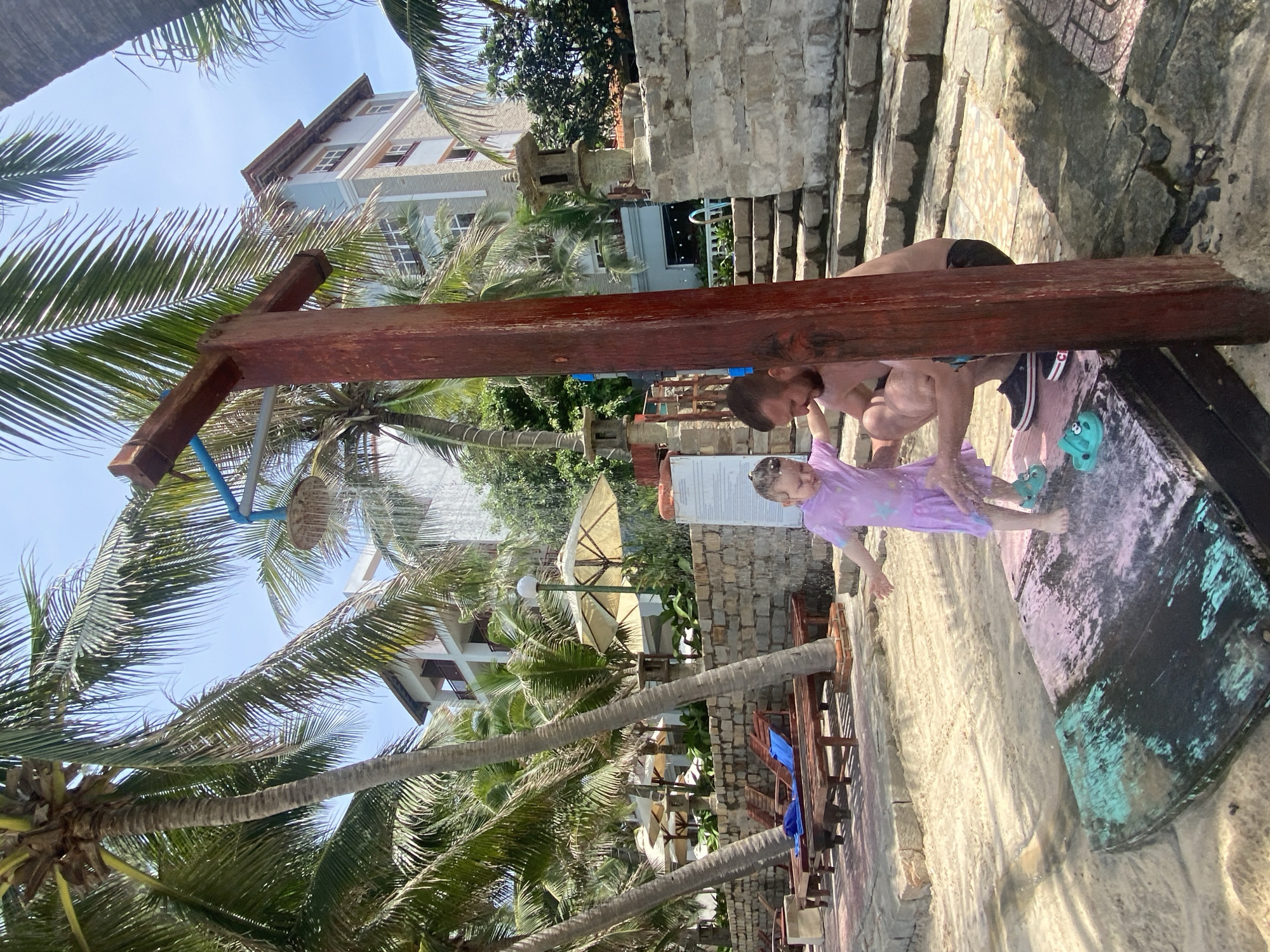 The image depicts a scene in Phu Thuy Ward, Vietnam, likely a resort or hotel area, with a focus on an outdoor shower. Two people, an adult and a child, are visible. The adult appears to be helping the child with the shower, as the child is standing under the spray. The child is wearing a purple outfit with a star pattern. The adult is wearing dark swimwear.
The setting is characterized by lush palm trees, a building in the background with multiple stories and windows, and a stone wall structure. There are also wooden benches and tables, suggesting an outdoor seating area. The ground is a mix of sand and concrete. The shower itself is constructed from wood, with a blue pipe leading to a showerhead.
The time of day appears to be daytime, given the bright sunlight casting shadows. The weather seems to be warm and pleasant, conducive to outdoor activities like showering. The overall mood is relaxed and suggests a casual, vacation-like atmosphere.
The image is oriented vertically, with the subjects and shower appearing towards the bottom and middle of the frame, while palm fronds and the building dominate the upper portions. The composition gives a unique perspective, looking up towards the sky through the palm trees.