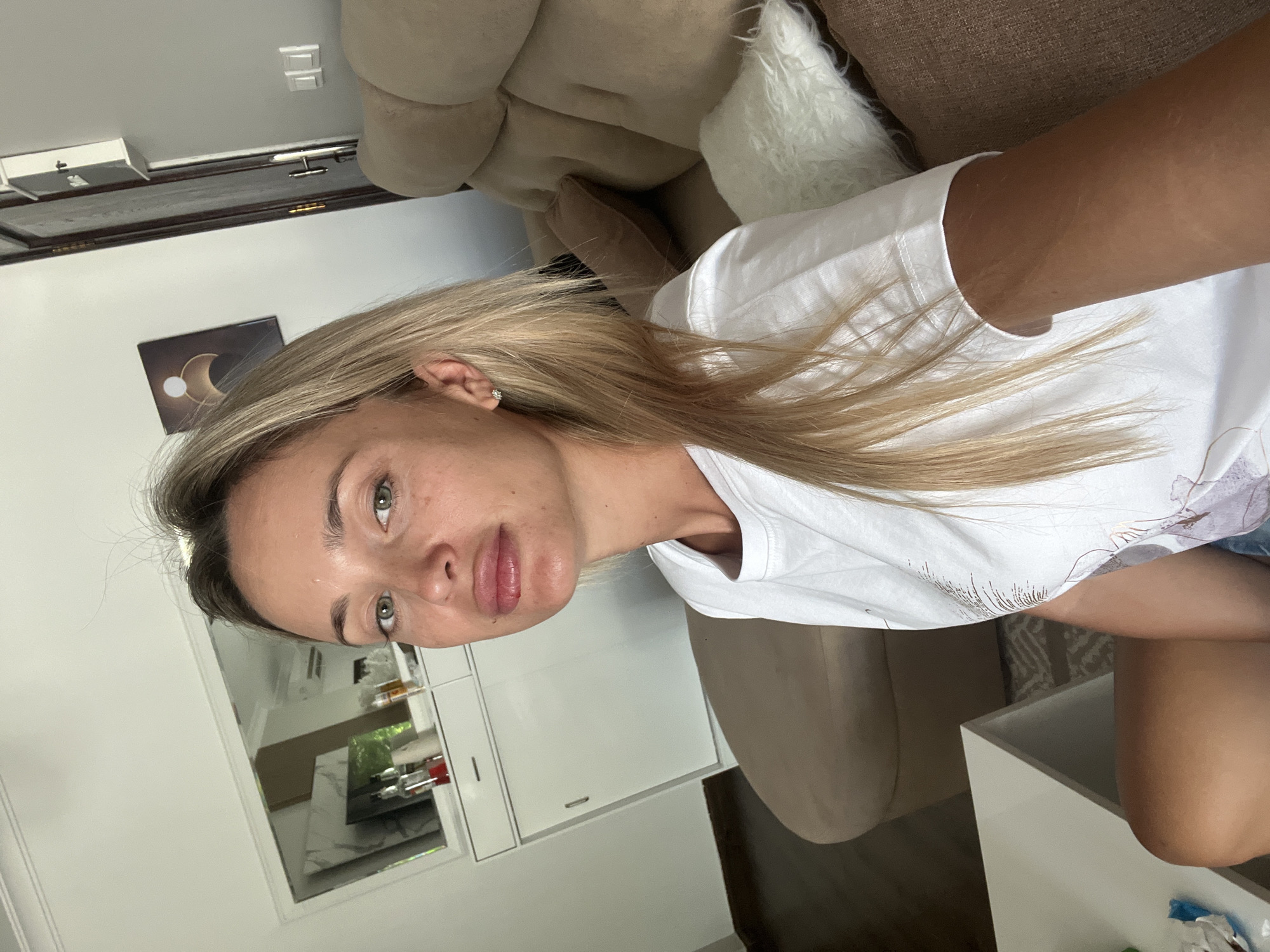 The image shows a young Caucasian woman with blonde hair and green eyes, taking a selfie while sitting on a couch. She is wearing a white t-shirt and appears to be in a well-lit room. In the background, there is a couch, a white cabinet with a mirror above it, and a door. The overall impression is of a casual, indoor setting, likely a living room or lounge area. The location is identified as Sharm El Sheikh, Egypt.