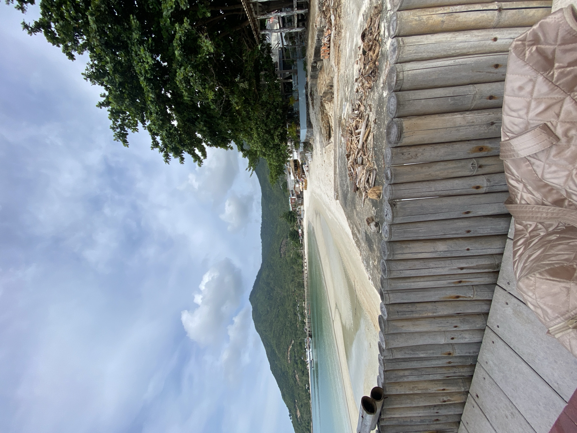 View from a bamboo-lined elevated position over the beach towards the mountains in Baan Chaloklum, Thailand. Clear sky with scattered clouds. Sandy beach with shallow water. Tree line along the beach. Buildings along the beach. Quilted jacket in the foreground.