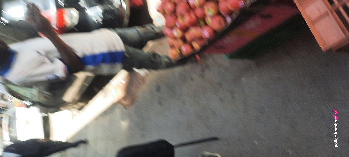 The image captures an outdoor street scene in Nairobi, Kenya, from an elevated vertical perspective. In the foreground on the right, a market display features a large quantity of red apples piled on a reddish surface, with a green crate visible directly below and a light brown wooden crate partially seen on the far right. Centered in the mid-ground, an individual with dark skin wears a white t-shirt with a vertical blue stripe and dark trousers. This person stands, holding a white smartphone in their right hand, their gaze directed towards the device. Behind this individual, a dark-colored vehicle, likely an SUV, is partially visible, either parked or slowly moving. Further in the background, another lighter-colored vehicle is discernible on a street. To the far left, a blurry figure dressed in a dark suit jacket and lighter trousers walks away from the camera. In the bottom-left corner, a black metal chair and the lower leg and foot of another person are partially visible. The ground surface is a dark, worn paved material. The text "police kombo 💕💕" is overlaid in white in the bottom-left corner of the image.