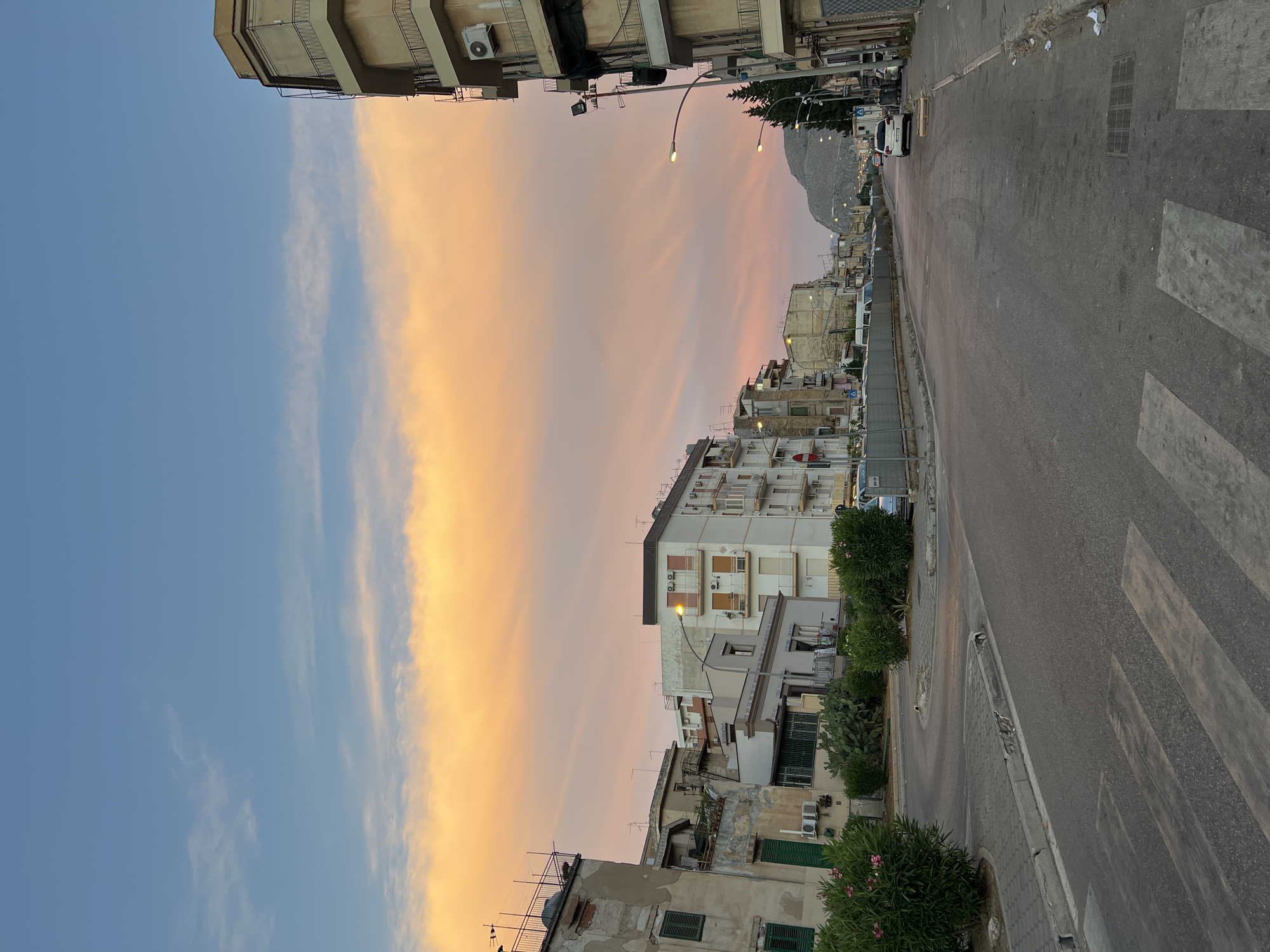 The image captures a street scene in Palermo, Italy, at dusk. The sky is a vibrant gradient of blue, orange, and pink, with wispy clouds stretching across. The foreground features a paved road with a pedestrian crossing. On the left side of the road, there are several buildings, some appearing aged and partially dilapidated, with green shutters and balconies. Lush green bushes with pink flowers are planted along the sidewalk. On the right side, a multistory building with balconies and air conditioning units is visible. Further down the street, more buildings line the road, and a mountain looms in the distance. A few cars are parked along the street and a street lamp illuminates the scene. The overall impression is of an urban environment transitioning into evening, with a dramatic sky as a backdrop.