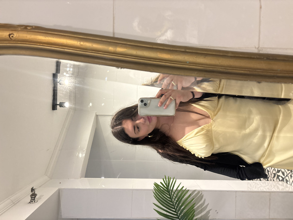The image depicts a female individual taking a mirror selfie within a tiled interior space in Petah Tikva, Israel. The subject, with long dark hair, is wearing a light yellow V-neck slip dress layered under a dark cardigan. She holds a light-colored smartphone with a clear case, visible in her reflection, angled towards the mirror to capture the image. Her visible right hand displays rings, and she wears gold-colored hoop earrings and a thin necklace. The background consists of white tiled walls with white crown molding near the ceiling. A ceiling-mounted track light fixture with two spotlights is reflected, and another fixture is present on the upper left wall. A green, palm-like decorative plant is situated on the left side of the reflection. The mirror features an ornate, gold-colored frame partially visible along its right edge. A section of black and white patterned floor tiles is present in the lower left of the frame.