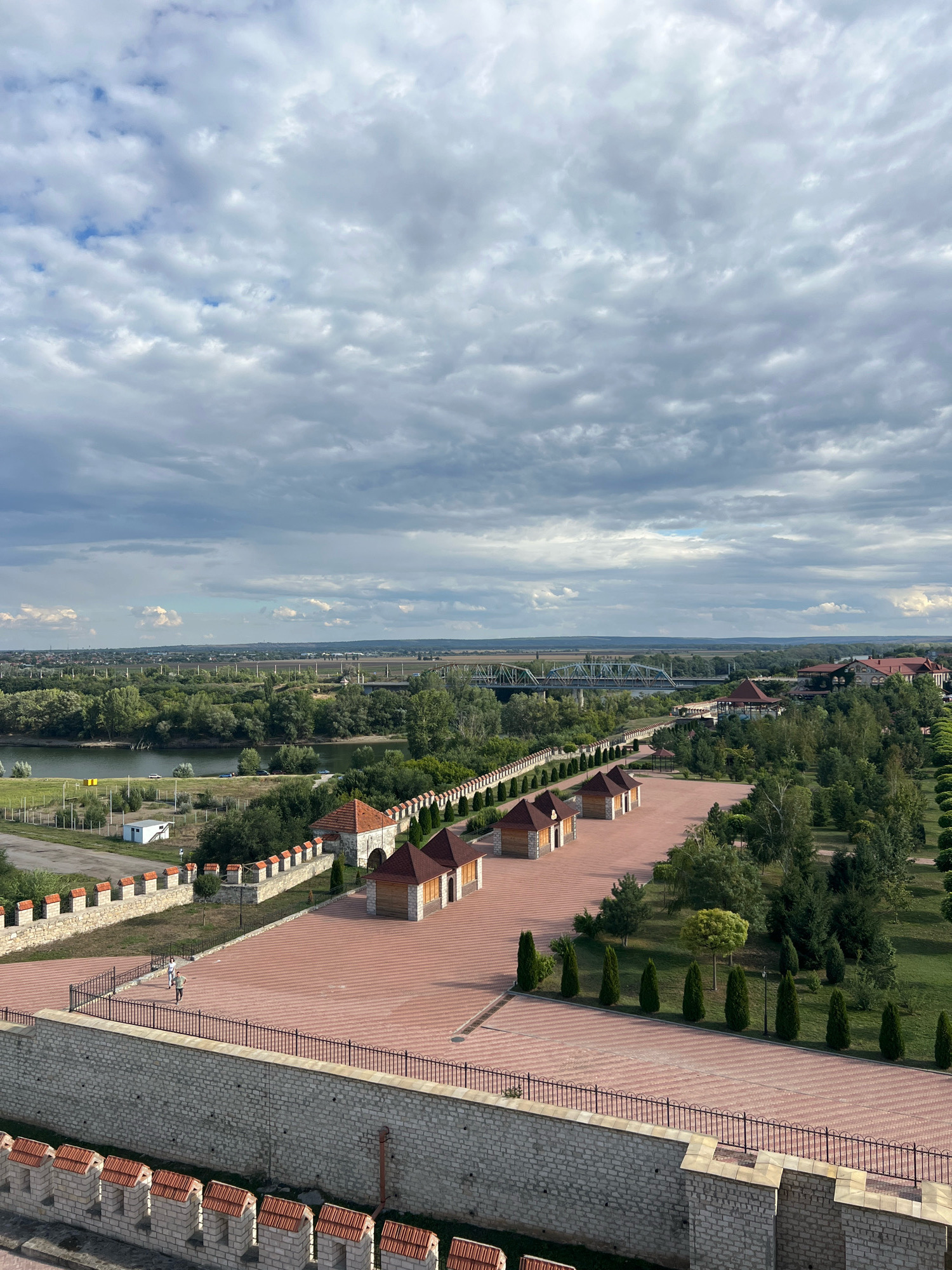 This image captures a scenic view of Bender Fortress in Bender, Moldova, under a cloudy sky. The fortress's stone walls, topped with red-tiled crenellations, form a prominent foreground. Beyond the walls, a red brick courtyard stretches out, lined with small, wooden structures featuring red-tiled roofs and neat rows of cypress trees. Further back, a river meanders through a landscape of lush green trees and vegetation. A bridge crosses the river in the distance, and more buildings, some with red roofs, are visible on the horizon, nestled amongst the trees. The sky is filled with dramatic, grey clouds, hinting at potential rain. Two individuals are seen walking on the courtyard, adding a sense of scale and life to the scene. The overall impression is one of historical architecture integrated with a beautiful natural environment.