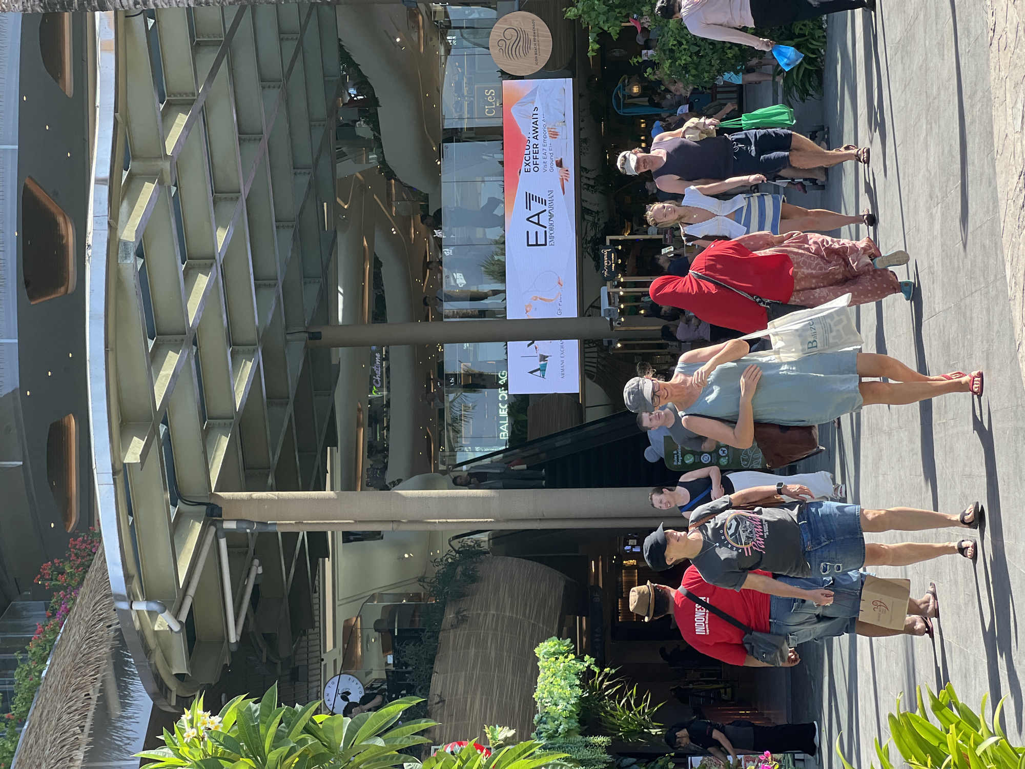 In a sunlit outdoor setting, likely in Denpasar, Indonesia, several people are seen walking and standing on a paved walkway in front of a modern building with a distinctive geometric facade. The time of day appears to be late morning or early afternoon, indicated by the strong shadows cast by the people.
The subjects include a diverse group of individuals, some in casual attire, suggesting a leisurely outing. One person in the foreground wears a red t-shirt with "INDONESIA" clearly visible on the back, and another person wears a t-shirt with the word "Paradis". Several women are also visible, one in a light blue dress and another in a white top with a striped skirt. Many are carrying shopping bags, including a prominent white bag labeled "Balize".
The building facade is composed of interlocking geometric shapes, creating a striking architectural element. Behind the people, large digital billboards display advertisements. One billboard features "EA7 EMPORIO ARMANI" with the text "EXCLUSIVE OFFER AWAITS". Another sign above the entrance reads "ICON BEACH PROMENADE", providing a strong clue to the location. The presence of "Chatime" also suggests a commercial or retail area.
The overall atmosphere seems relaxed and bright, characteristic of a sunny day in a tropical or subtropical region. The people appear to be enjoying their time, either shopping or simply passing through the area. The combination of modern architecture, retail advertisements, and casual pedestrians paints a picture of a bustling commercial and leisure hub.