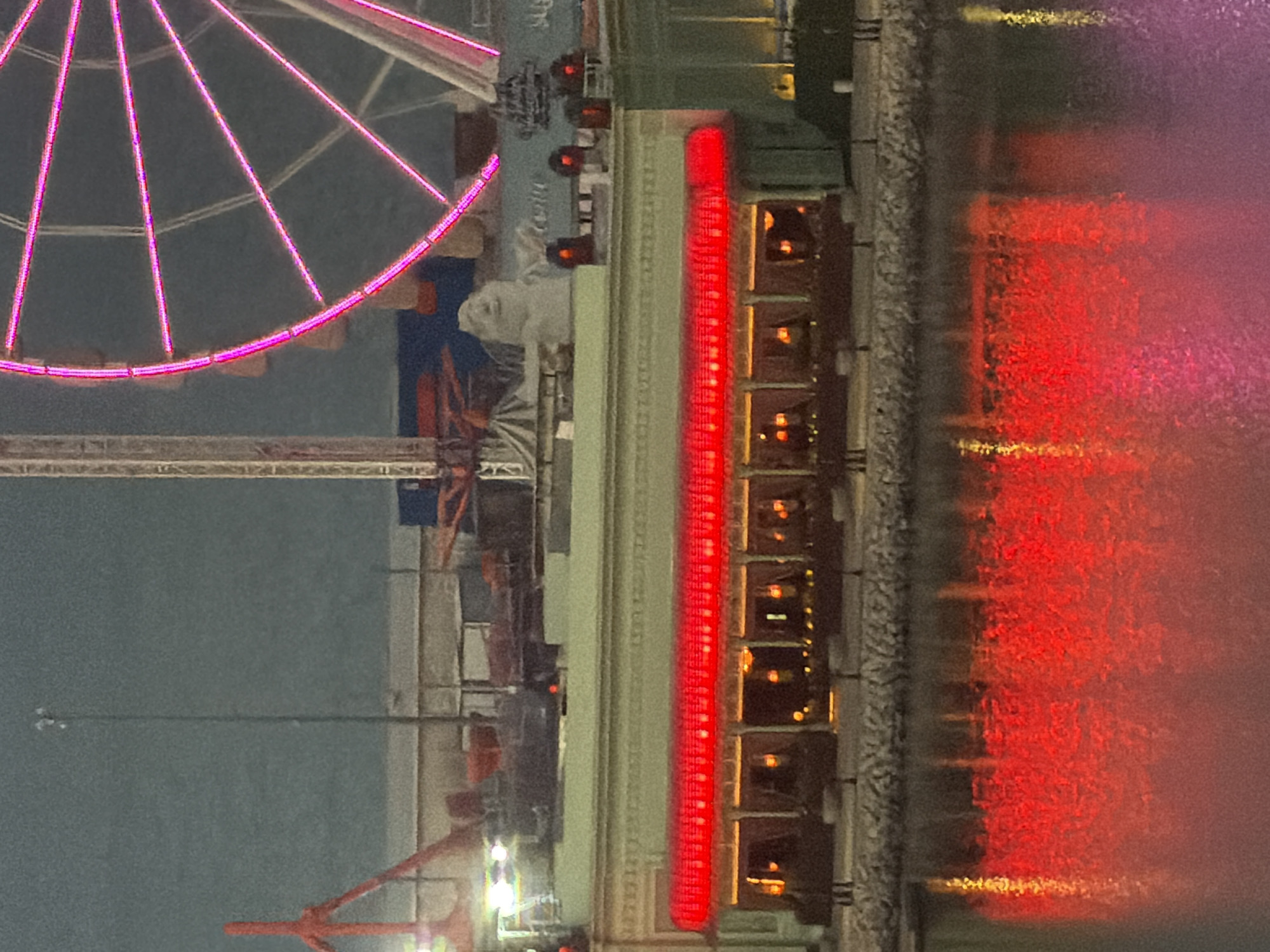 The image depicts a waterfront scene at dusk or night. In the foreground, a body of water reflects the bright red neon lights of a building. The building itself has a facade with awnings and windows, behind which warm interior lights are visible. Above the building, a Ferris wheel with pink lights is prominently featured, along with a tall structure that appears to be part of a carnival ride or construction crane. The sky is a muted grey, suggesting overcast conditions or twilight. The overall impression is of an amusement park or entertainment district by the water.