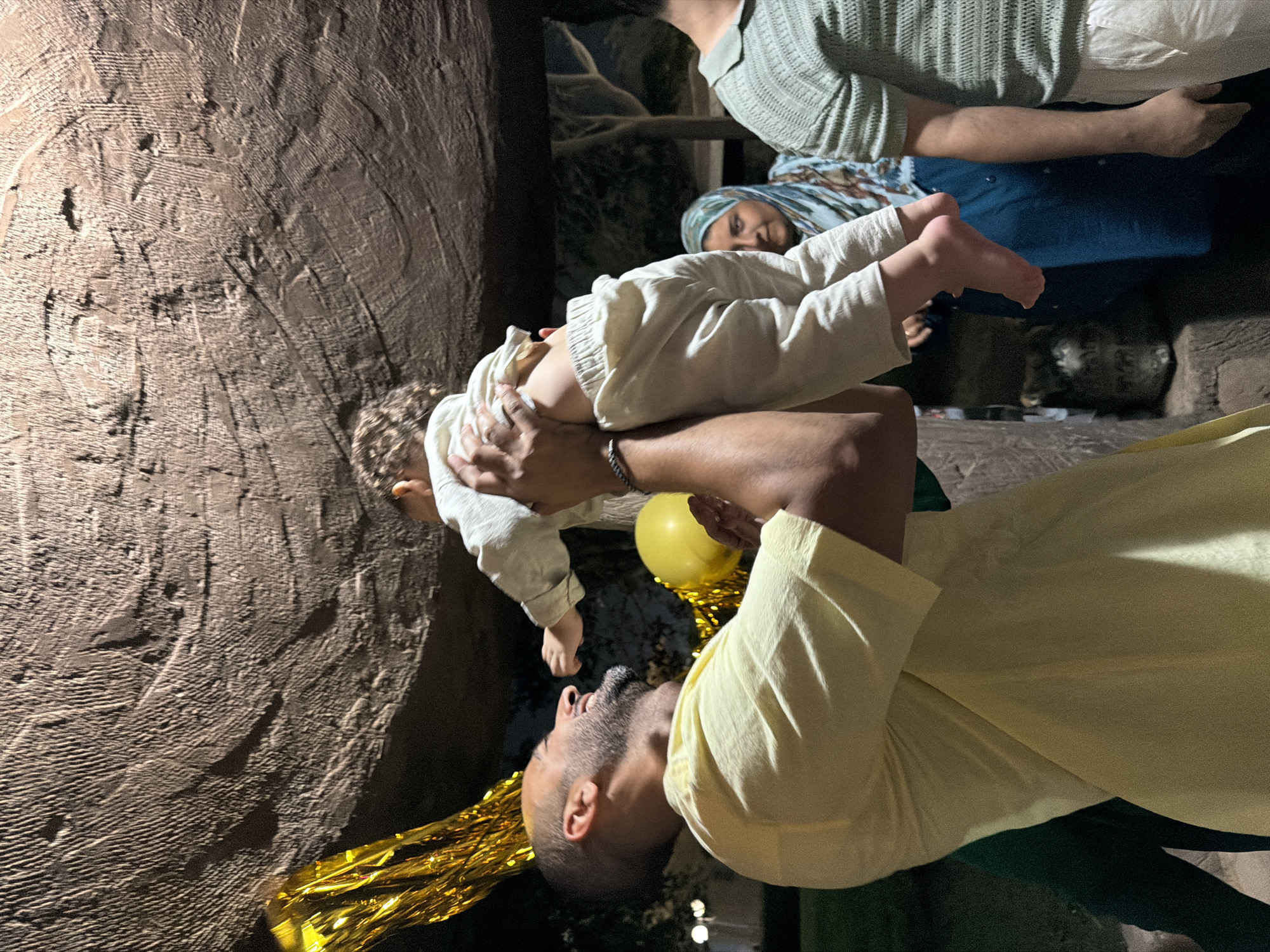 A man in a yellow shirt is holding a baby up in the air. The baby is wearing light-colored clothing. There is a yellow balloon and some gold streamers in the background. Behind them, a woman wearing a hijab and a person in a green striped shirt are visible. The scene appears to be outdoors, with trees and artificial lighting visible in the background, suggesting an event or gathering. The rough, textured ceiling above suggests an indoor-outdoor space, possibly a cafe or restaurant with an unusual architectural feature, consistent with some modern designs in New Cairo or 6th of October City, Egypt.