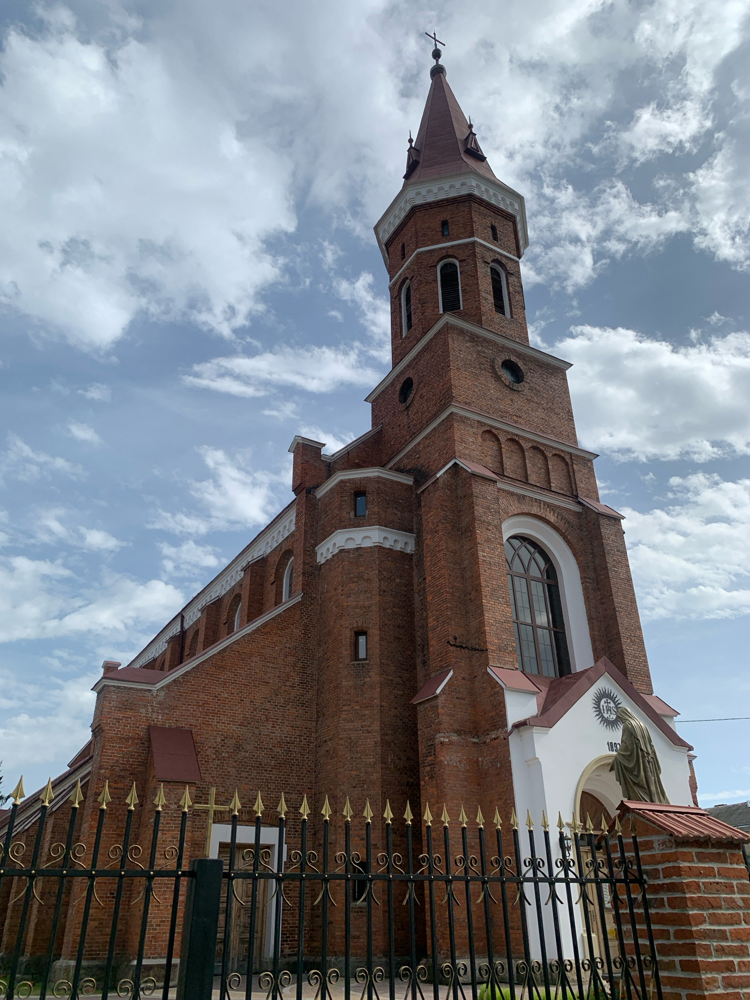 The image shows a brick church with a tall, octagonal bell tower topped by a red spire with a cross. The church has arched windows and decorative white trim. In front of the church is a black metal fence with gold finials. A statue of a robed figure stands near the entrance. The church is located in Kolomyia, Ukraine. The sky is cloudy.