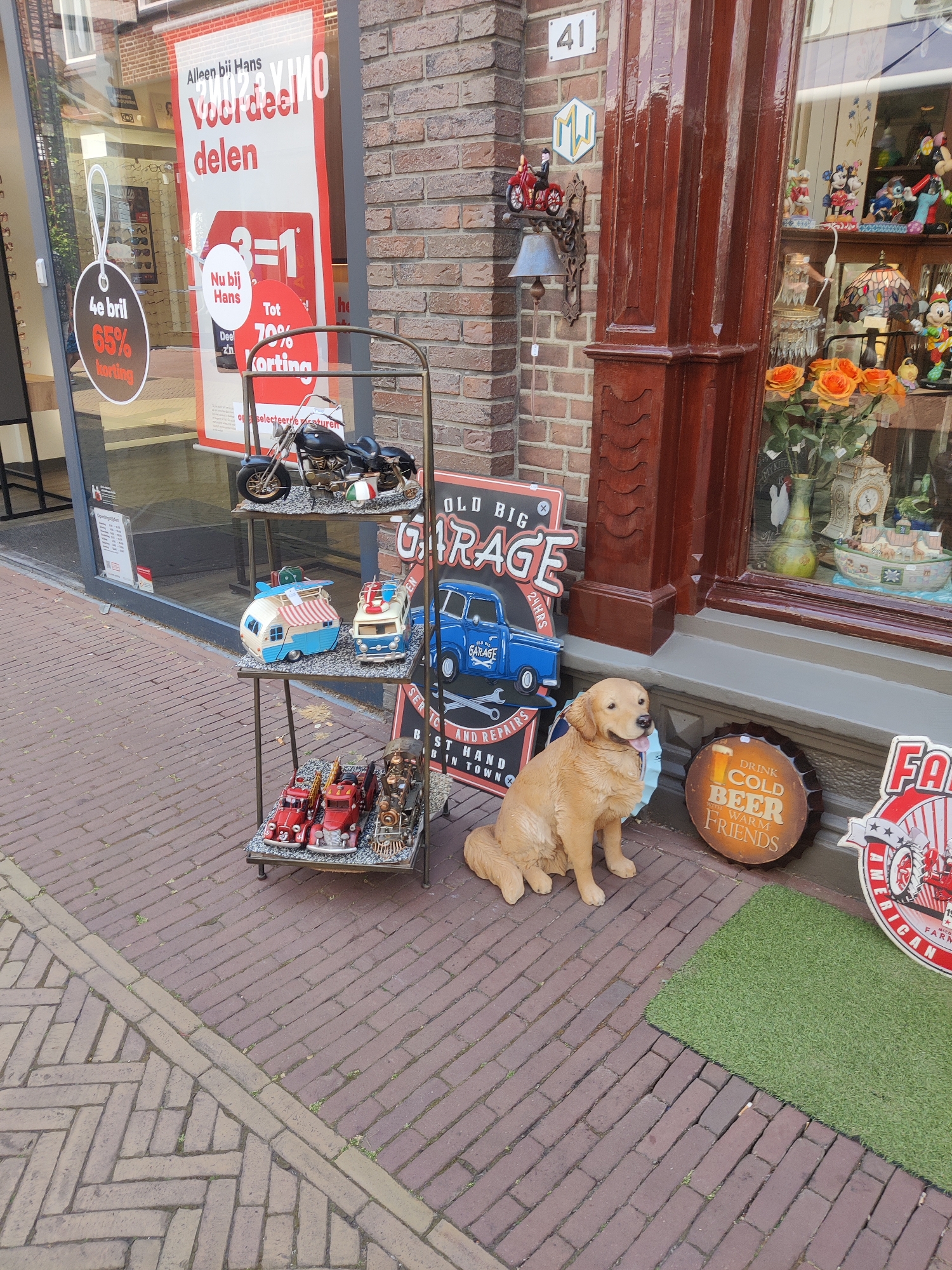 A golden retriever dog is sitting on a brick sidewalk outside a storefront. The dog is facing left with its tongue hanging out. To the right of the dog is a round metal sign with the words "DRINK COLD BEER WITH WARM FRIENDS" and an image of a beer. To the left of the dog is a metal shelf with several vintage toy vehicles, including a motorcycle, a camper, a fire truck, and a train. Behind the shelf is a large red and white advertisement banner for a sale. The storefront has a large glass window displaying various items, including a stained-glass lamp and figurines. A brick building facade is visible above the storefront. The sidewalk is made of red brick pavers arranged in a herringbone pattern. The overall scene suggests an outdoor display for a shop. The location is Winterswijk, Netherlands, as indicated by the presence of the number 41 on the building, which is a common house number.