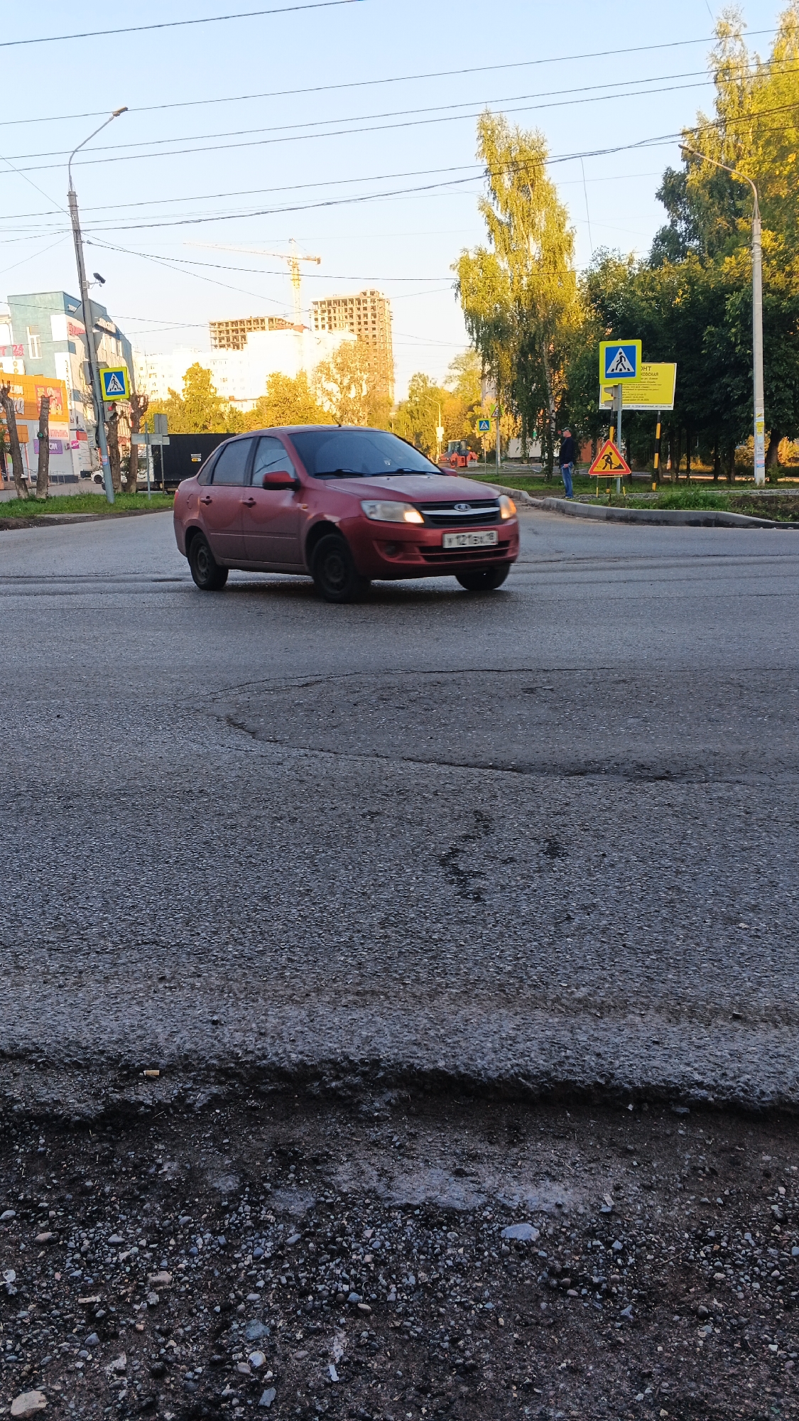 A red sedan, a Lada Granta, with license plate Y121BX 18, is driving on a paved road. The car is in the middle of a road intersection. In the background, there are trees, buildings under construction, a street light, and traffic signs. One sign indicates a pedestrian crossing, and another sign shows a warning for a pedestrian. There is one person standing on the side of the road near the pedestrian crossing sign. The road surface in the foreground is rough and appears to have potholes. The scene is set in the city of Izhevsk, Russia, as indicated by the license plate. The lighting suggests it is either early morning or late afternoon.