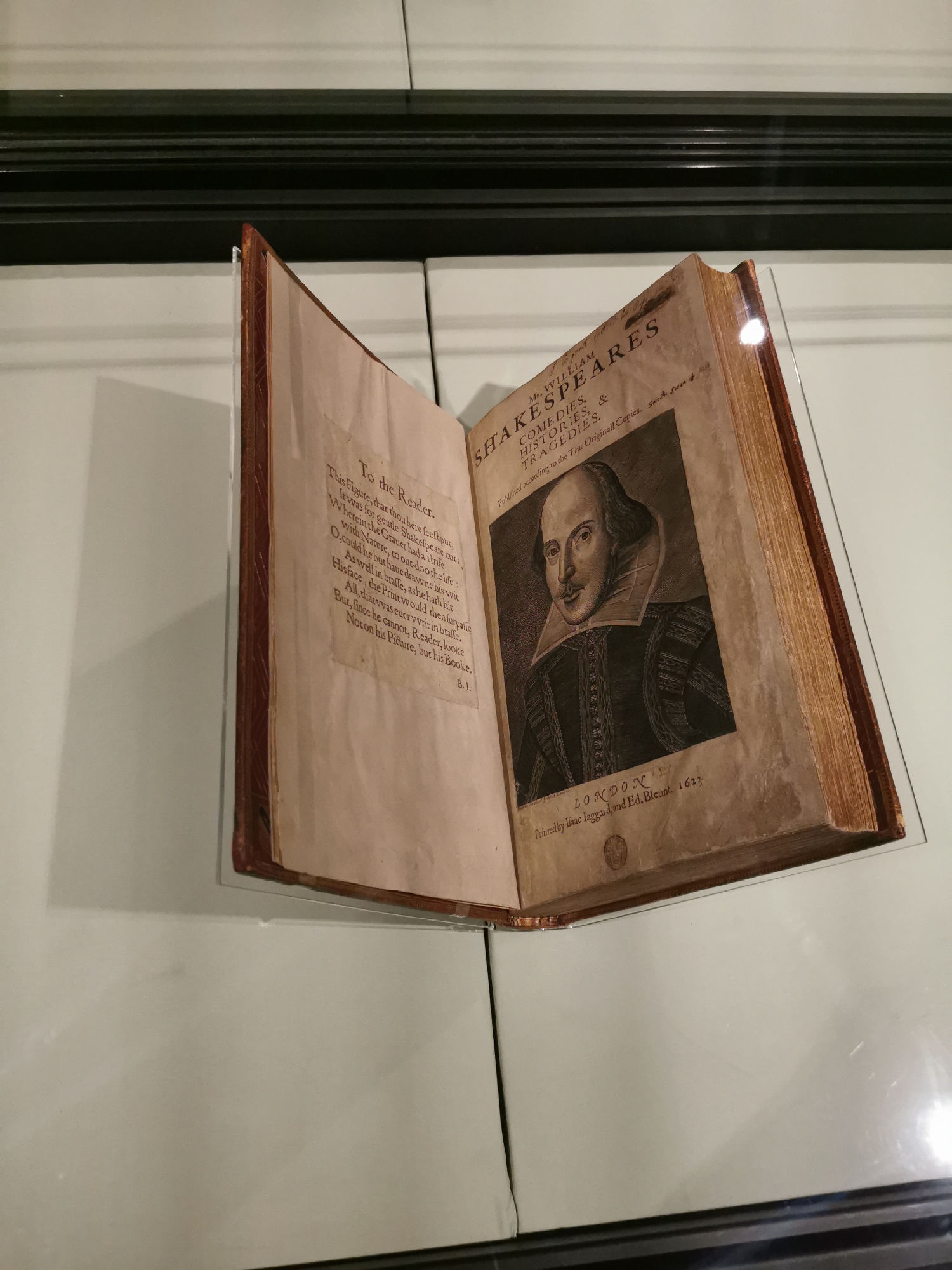 An open, aged book, identified as a copy of "Mr. WILLIAM SHAKESPEARES COMEDIES, HISTORIES, & TRAGEDIES," printed by Isaac Iaggard and Ed. Blount in London in 1623, is displayed on a transparent stand. This is the First Folio, the first collected edition of Shakespeare's plays.
The right page features a prominent engraved portrait of William Shakespeare, depicted with a receding hairline, a small beard and mustache, and a large ruff collar. Above the portrait, the title and publication details are visible. The left page presents a poem titled "To the Reader." The first lines of the poem read: "This Figure, that thou here seest put, / It was for gentle Shakespeare cut; / Wherein the Graver had a strife / With Nature, to out-doo the life." The poem concludes, "But, since he cannot, Reader, looke / Not on his Picture, but his Booke."
The book's pages exhibit significant yellowing and age-related discoloration, with the fore-edge appearing gilded and worn. The item is protected by a clear acrylic or glass display case, indicating its value as an artifact. The setting is indoors, likely an exhibition space such as a museum or library, with a neutral-toned background and a dark, horizontal molding above the book. A bright reflection is visible on the right edge of the book, suggesting focused lighting for display. The overall scene suggests careful preservation and presentation of this historic literary work.