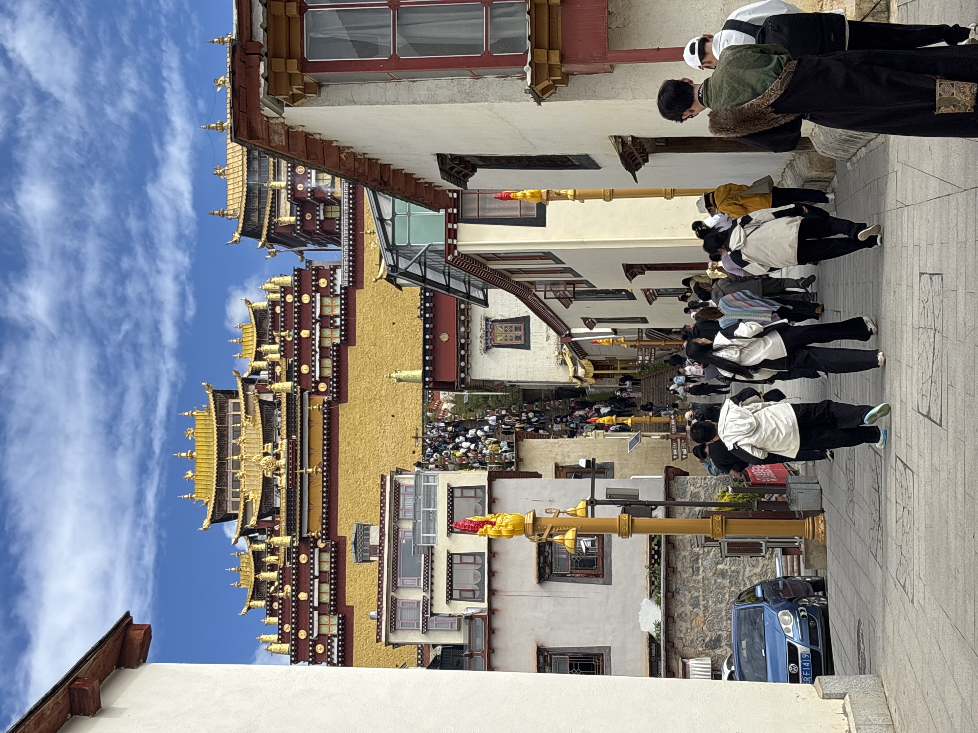 This is a medium shot, eye-level, outdoor, vertical photograph taken on a sunny day in Shangri-La, China. The image depicts a street scene with a large Tibetan monastery complex prominently featured in the background. The monastery has golden roofs and intricate red and white detailing. A crowd of tourists and locals are walking up a long set of stairs towards the monastery. In the foreground, several people are walking on a paved street. A blue Volkswagen car is parked on the left side of the street. The buildings on either side of the street are white with red trim. The sky is blue with scattered white clouds. The overall atmosphere is one of a popular tourist destination with significant cultural and religious significance.