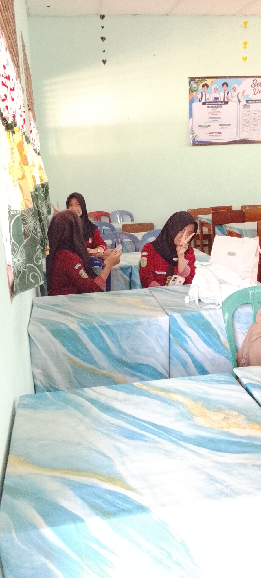 Three young women wearing maroon shirts and black headscarves are seated at tables in what appears to be a classroom or common area. The room has light blue walls and white ceiling tiles. A poster with student portraits and organizational charts is displayed on the wall. Heart-shaped decorations hang from the ceiling. The tables are covered with a blue and white marble-patterned cloth. One woman in the foreground is holding a cell phone and making a peace sign. Another woman to her left is looking at her phone. The third woman, further back and to the right, is also looking at her phone and is partially obscured by the table and a white bag. The scene suggests a casual gathering or study session in an Indonesian educational institution, possibly in Jepara, a city known for its educational facilities.