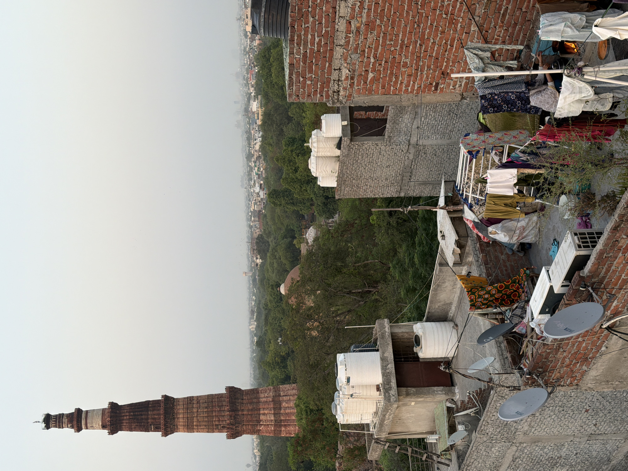 This is a medium shot, taken from a high vantage point, of the Qutb Minar in Delhi, India, with a residential rooftop in the foreground. The Qutb Minar, a UNESCO World Heritage site, is a tall, red sandstone tower with intricate carvings and several balconies. It is located on the left side of the image. In the middle ground, there is dense greenery and some domed structures, suggesting other historical buildings within the complex.
The foreground is dominated by the roofs of residential buildings. Several large white water tanks are visible on the flat roofs, a common sight in Indian cities. On one rooftop, there is a clothesline with various garments hanging to dry, including colorful patterned fabric and plain clothing. A woman is visible in the lower right corner, near a fire pit, tending to something, possibly cooking. Several satellite dishes are mounted on walls. The environment appears somewhat hazy, typical of urban areas, with a distant cityscape visible on the horizon. The overall scene depicts a juxtaposition of ancient history and modern daily life in Delhi.