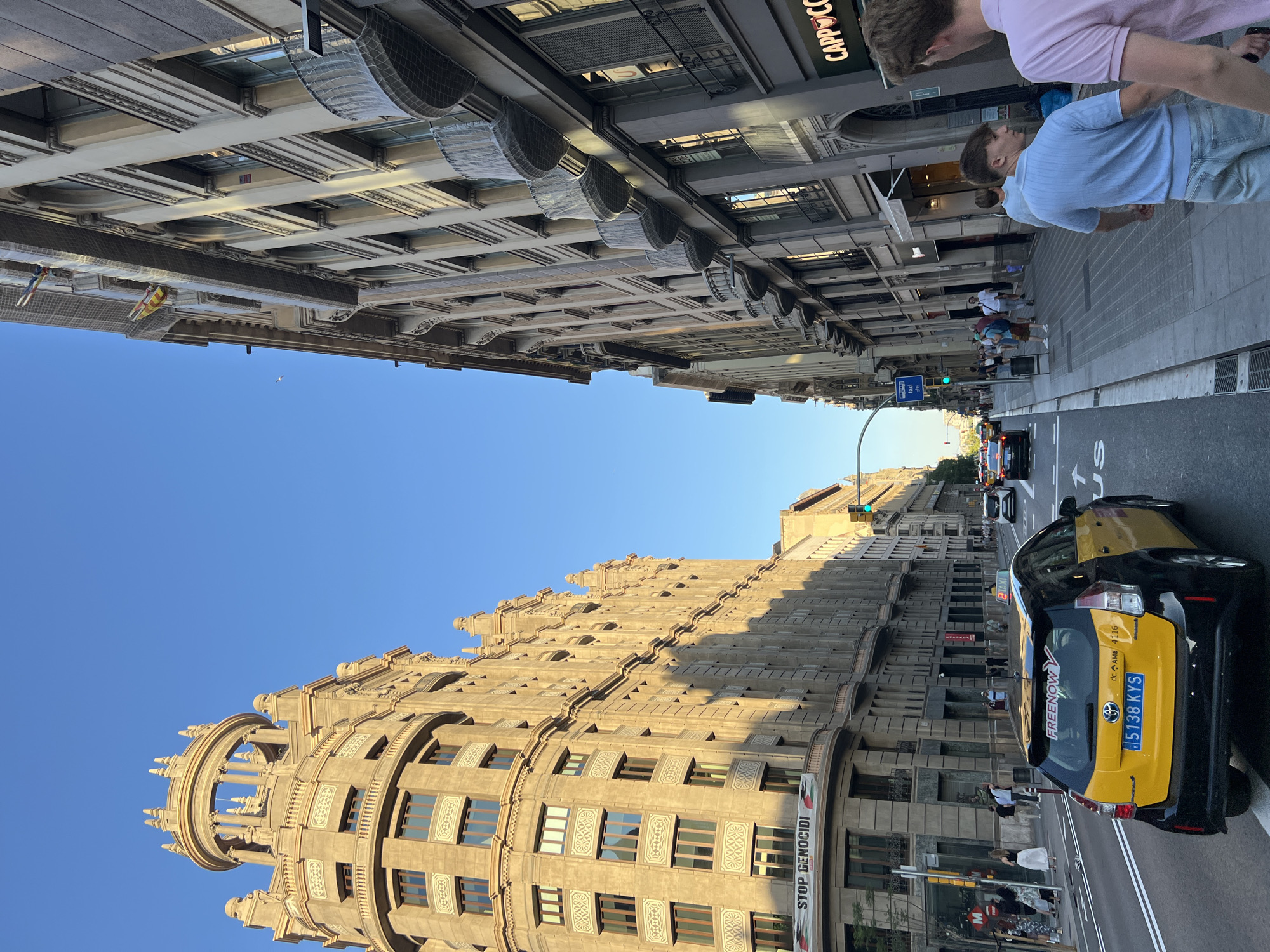The image captures a street scene in Barcelona, Spain, bathed in the warm light of a late afternoon or early morning sun. The perspective is from a vehicle on a multi-lane road, looking down a wide avenue flanked by ornate, historic buildings.
On the left, a prominent, cream-colored building features a distinctive circular turret with arched windows and decorative elements. Its facade is adorned with repeating patterns and numerous windows. A banner with the text "STOP GENOCIDI" is visible on a lower section of this building.
The right side of the street is dominated by a taller, more densely built apartment or office building with a light-colored facade. It has a rhythmic pattern of windows and balconies, many of which are covered with netting. Signs for businesses, including "CAPPPUCCINO," are visible at street level.
In the foreground, a yellow and black taxi with a "FREEnOW" sticker on its rear window is prominent. Its license plate reads "5138 KYS". Several other vehicles are present on the road, moving in the same direction.
On the sidewalks, people are walking, some singly and others in small groups. Their attire suggests casual summer clothing. The environment is urban, with streetlights, traffic signals, and a clear blue sky overhead. The overall impression is a bustling city street with a blend of architectural styles and everyday life.