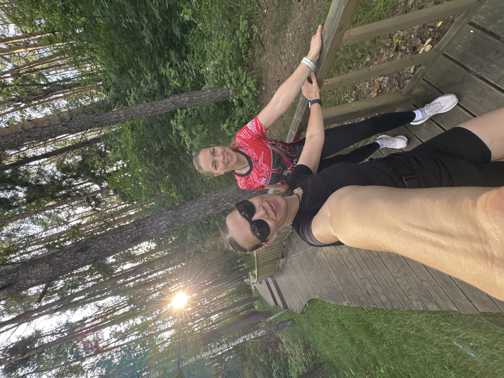 Two women are taking a selfie on a wooden boardwalk in a forest. The woman in the foreground is wearing sunglasses and a black t-shirt and shorts. The woman in the background is wearing a red t-shirt and black leggings. Both women are smiling. The boardwalk appears to be made of weathered wood and curves to the left. The forest is dense with tall trees and green foliage. The sun is setting, casting long shadows and lens flare across the image. The scene is likely located within the city of Chaikovsky, Russia, given the presence of a forest and boardwalk, which are common recreational features in the area.