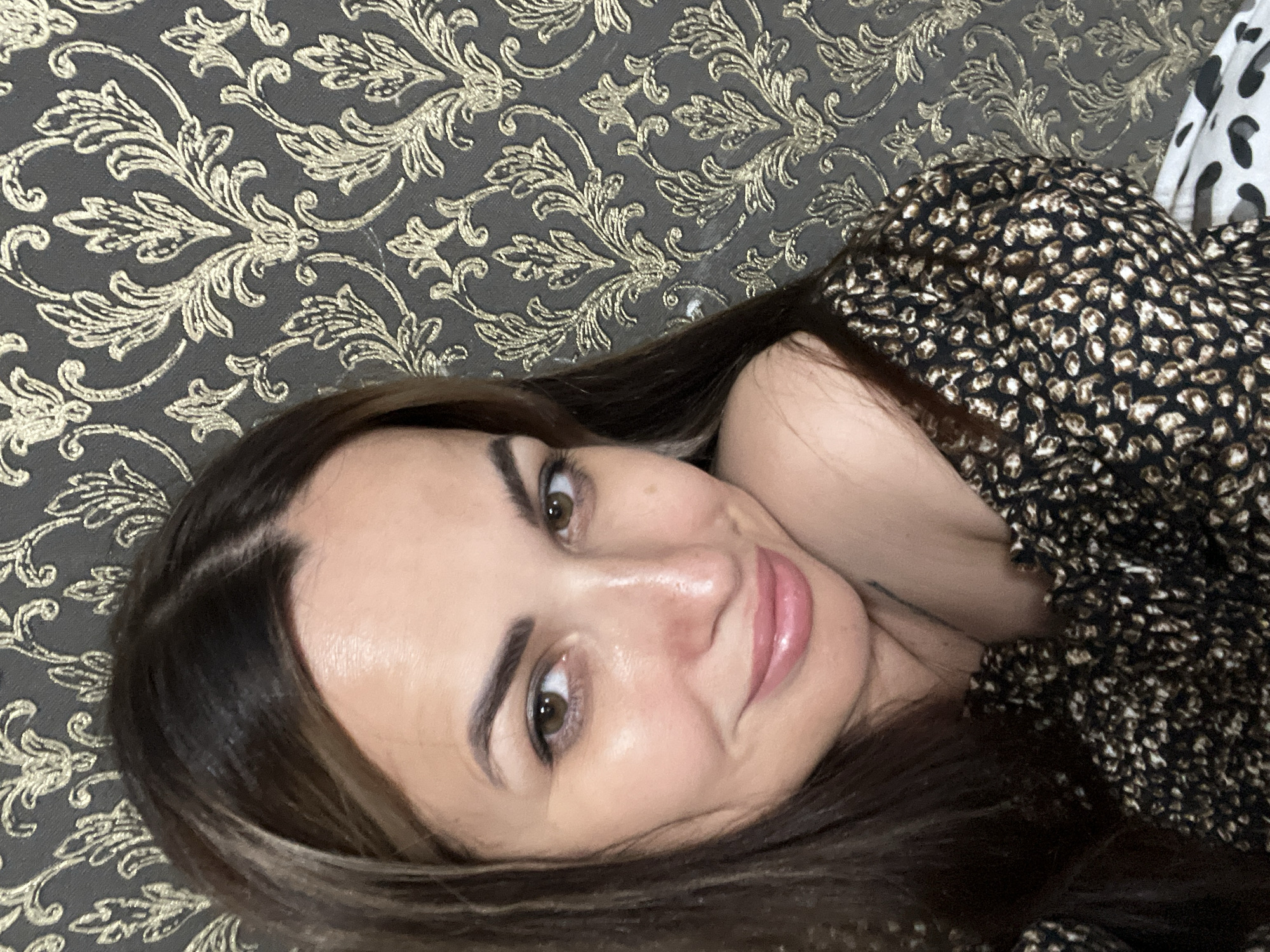 A medium shot photo shows a woman from the shoulders up, posed in front of a grey patterned wallpaper. The woman has long, dark brown hair parted in the middle and falling over her shoulders. Her skin is fair and she is wearing makeup, including lip gloss. Her eyes are brown and she is looking towards the camera with a slight smile. She is wearing an off-the-shoulder top with a black and brown animal print pattern. The wallpaper behind her features a gold damask pattern. The lighting suggests the photo was taken indoors, possibly in a home setting.