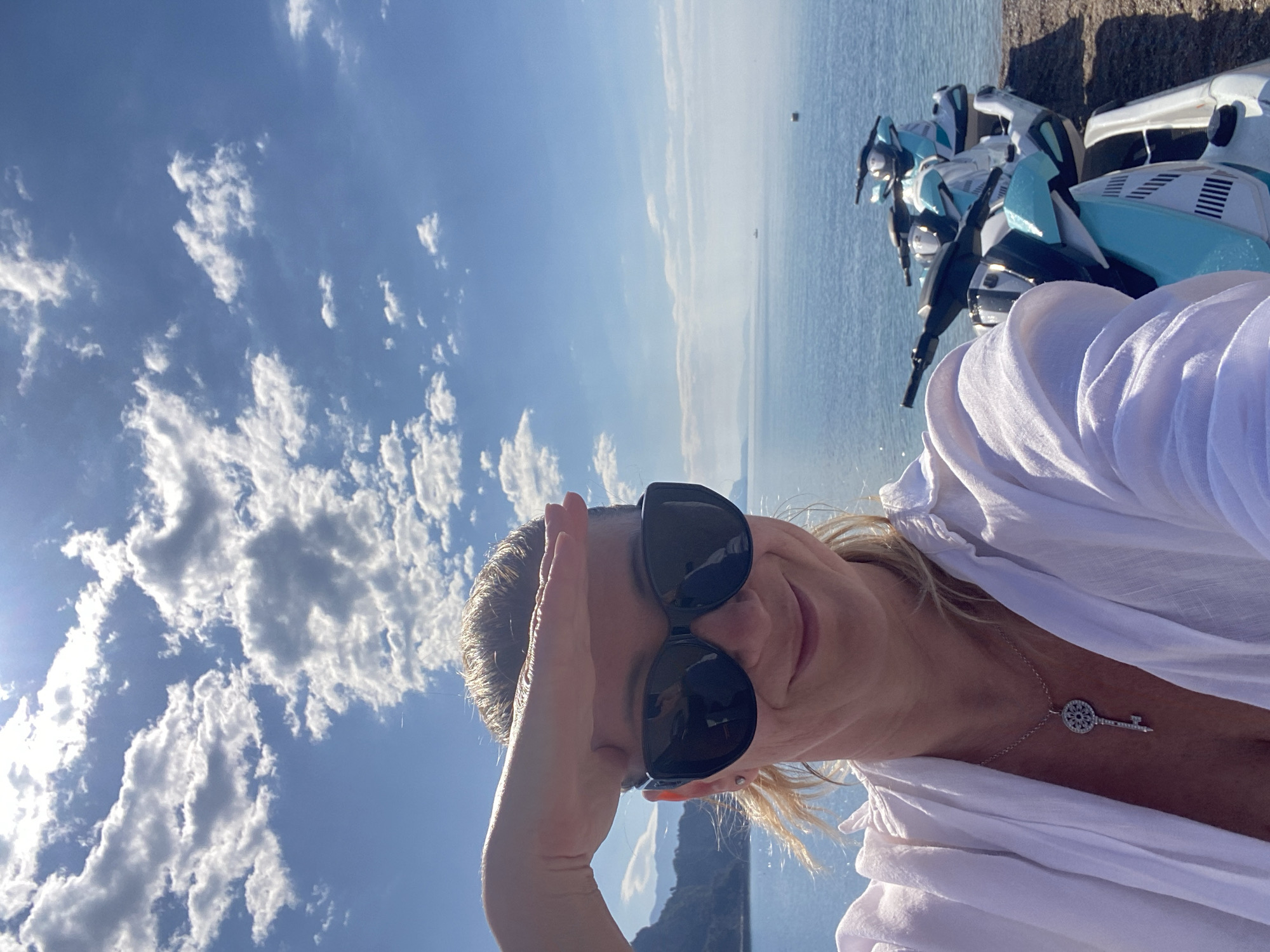 The image depicts a woman with blonde hair and sunglasses, shielding her eyes with her hand. She is wearing a white shirt and a silver necklace with a key pendant. The background shows a body of water, presumably the sea, with a sky full of scattered clouds. In the upper right corner of the image, there are several blue and white jet skis. The photo appears to be taken at a high angle, with the horizon tilted. The location, Prijevor, Montenegro, is inferred from the presence of the sea and coastline.