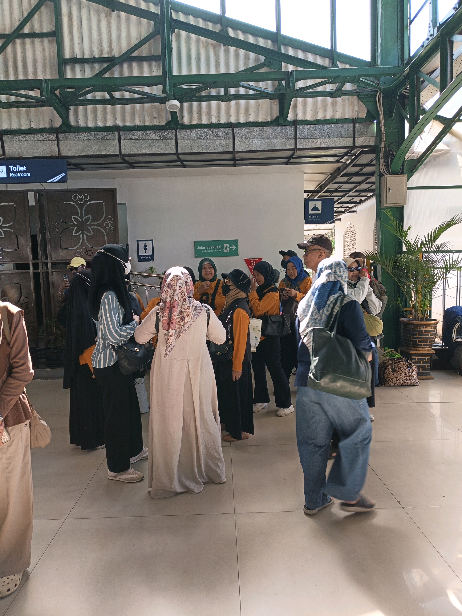 In the city of Surakarta, Indonesia, a group of people, mostly women in hijabs, stand on a tiled floor within a covered area that appears to be a public transportation hub, possibly a train station, given the overhead structure of green metal beams and corrugated roofing. A blue sign with a wheelchair symbol and the word "Toilet" is visible. Another sign in green indicates "Jalur Evakuasi" (Evacuation Route). Several individuals are dressed in traditional Muslim attire, including long dresses and hijabs in various colors and patterns. Some are holding bags, and one man in the background wears a baseball cap. The overall impression is a scene of people waiting or passing through a functional, open-air public space.