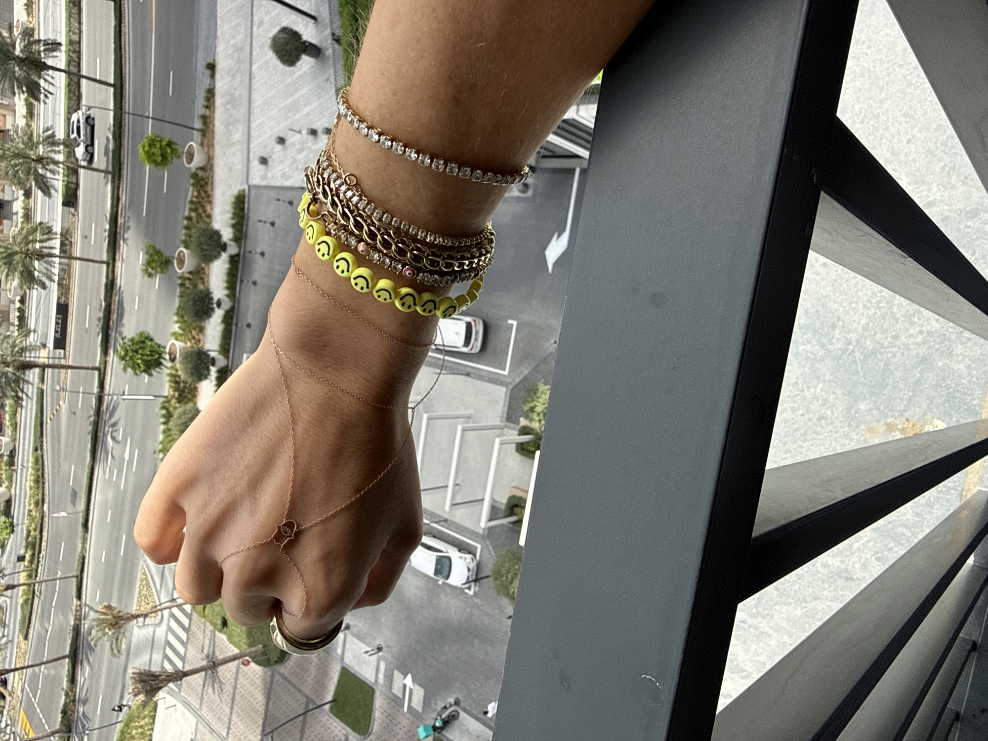 A high-angle, medium-shot captures a person's left arm and hand adorned with multiple bracelets and a ring, extending over what appears to be a balcony railing. The wrist wears several gold and yellow beaded bracelets, including one featuring smiley face designs, along with a sparkling bracelet. The hand has a thin, decorative chain-like piece of jewelry connected to a ring on the index finger. The backdrop reveals a cityscape, specifically a neighborhood in Dubai, United Arab Emirates, with roads, palm trees, buildings, and parked cars visible below. The perspective is from an elevated position, suggesting the photograph was taken from a high-rise building.