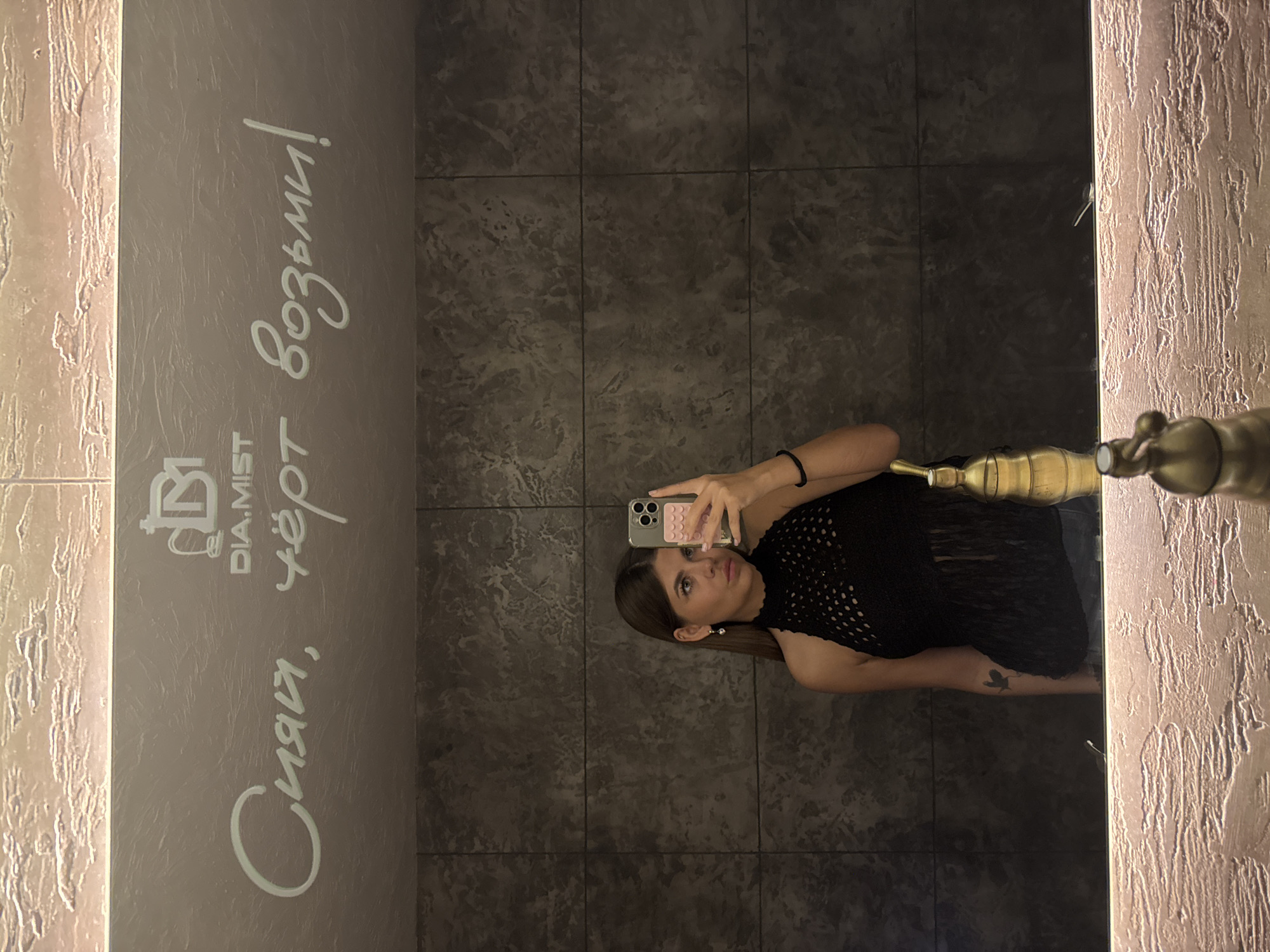 A young woman with shoulder-length brown hair takes a mirror selfie in a dimly lit restroom. She is wearing a black crocheted halter top with fringe detailing and denim shorts. She is looking into the mirror, holding up her iPhone with a pink case in her right hand. She is wearing a heart-shaped earring in her left ear. To her right, in the mirror's reflection, is a gold-colored faucet. Above her, on the mirror, is a logo for "DIA.MIST" and white text in Cyrillic script that reads "Сияй, чёрт возьми!" which translates to "Shine, damn it!". The restroom walls are made of dark grey textured tiles, and the mirror is framed by a textured material with embedded lights. The overall scene suggests a fashionable establishment. It is not possible to determine the exact location within Chaikovsky, Russia based on the provided image.