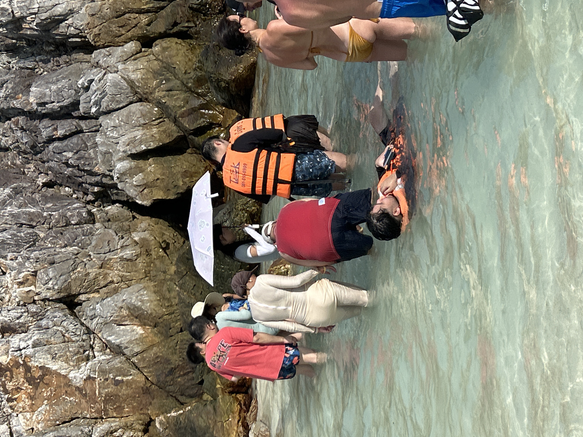 The image shows a group of people in shallow, clear turquoise water near a rocky shore. The location is Phru Nai, Thailand. Several individuals are wearing life vests, some orange and some black and white striped. One person is lying in the water, appearing relaxed, wearing an orange life vest. Another person in a red shirt and blue patterned shorts is walking through the water. A person in a full-length beige swimsuit is also wading. In the background, a large rock face rises from the water. A white umbrella is held by someone standing in the water. Some people are facing away from the camera, while others are partially visible. The water is calm, with sunlight reflecting off its surface.