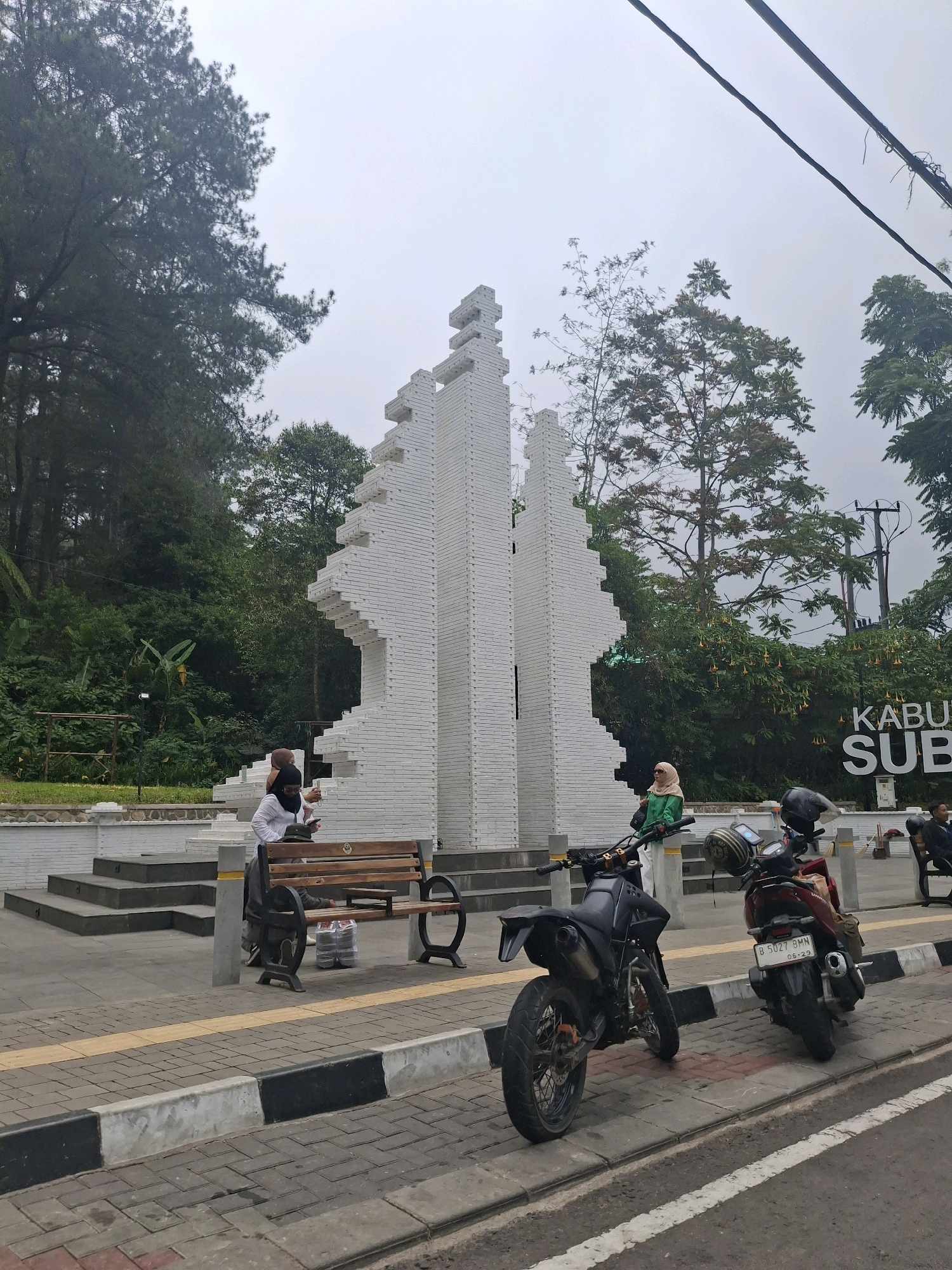 The image depicts a public space in Subang, Indonesia, featuring a unique white brick monument with a tiered, stepped design. To the left of the monument is a lush green hillside covered in trees. In front of the monument, a wooden bench is occupied by two individuals. One person, wearing a white top and a dark hijab, is holding a phone. Another person, wearing a dark top and hijab, is also seated. A third person, dressed in a green jacket and light-colored pants, stands near a black motorcycle parked on the sidewalk. Another motorcycle, a red scooter, is parked to the right of the black motorcycle. A sign in the background reads "KABUPATEN SUBANG". The ground is paved with interlocking bricks, and there are yellow and black-striped curbs along the road. The sky is overcast.