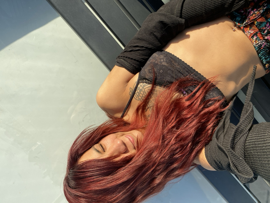 A woman with red hair is lying down wearing a black lace bra top, floral shorts, and a black, ribbed long-sleeve shirt. She has a tattoo on her shoulder. Her head is tilted towards the camera as she smiles. The scene is bathed in sunlight. In the background, there's a black-and-white structure, possibly a fence or a set of blinds.