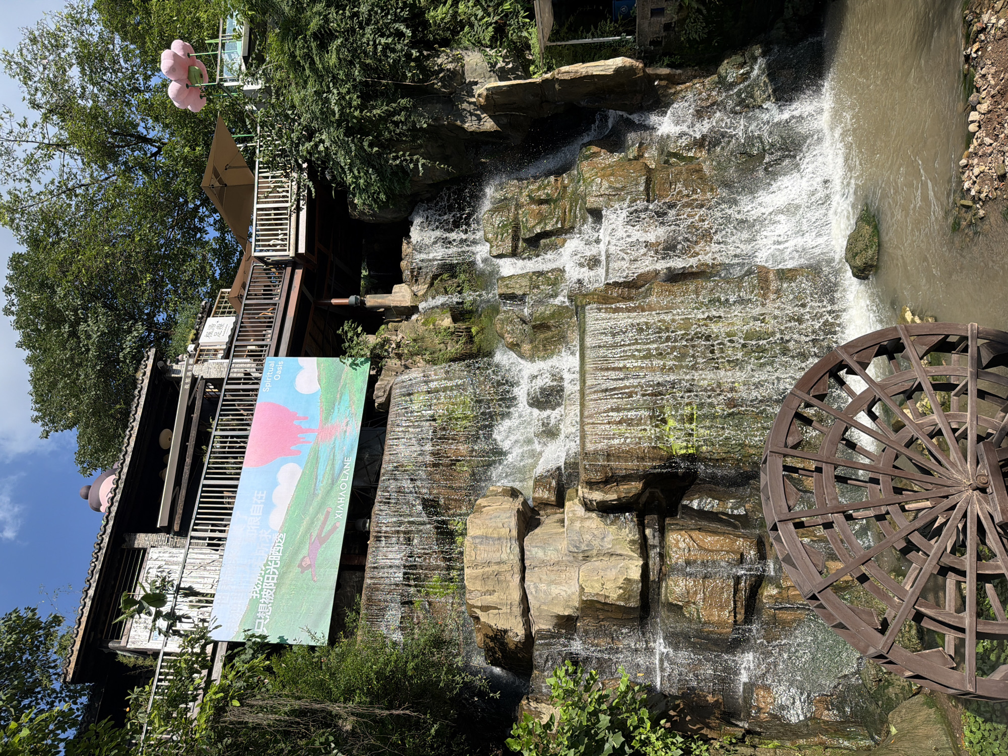 The image captures a scenic outdoor view of a multi-tiered waterfall cascading over rocks, with a wooden water wheel prominently positioned in the foreground at the base of the waterfall. Above the waterfall, a building with traditional Chinese architecture is visible, featuring a balcony or walkway with white railings. A large banner displaying a cartoonish image of a person lying on a grassy hill with a pink, dripping shape and text is hung on the side of the building. Lush green trees and vegetation surround the scene, and the sky above is blue with some clouds. In the upper right portion of the image, a cluster of pink, balloon-like objects are suspended. This scene is located in the Nan'an District of China, likely a tourist attraction or a place of leisure. No people are clearly discernible in the image, and the water wheel is stationary. The overall impression is one of a tranquil, natural setting enhanced by man-made structures.