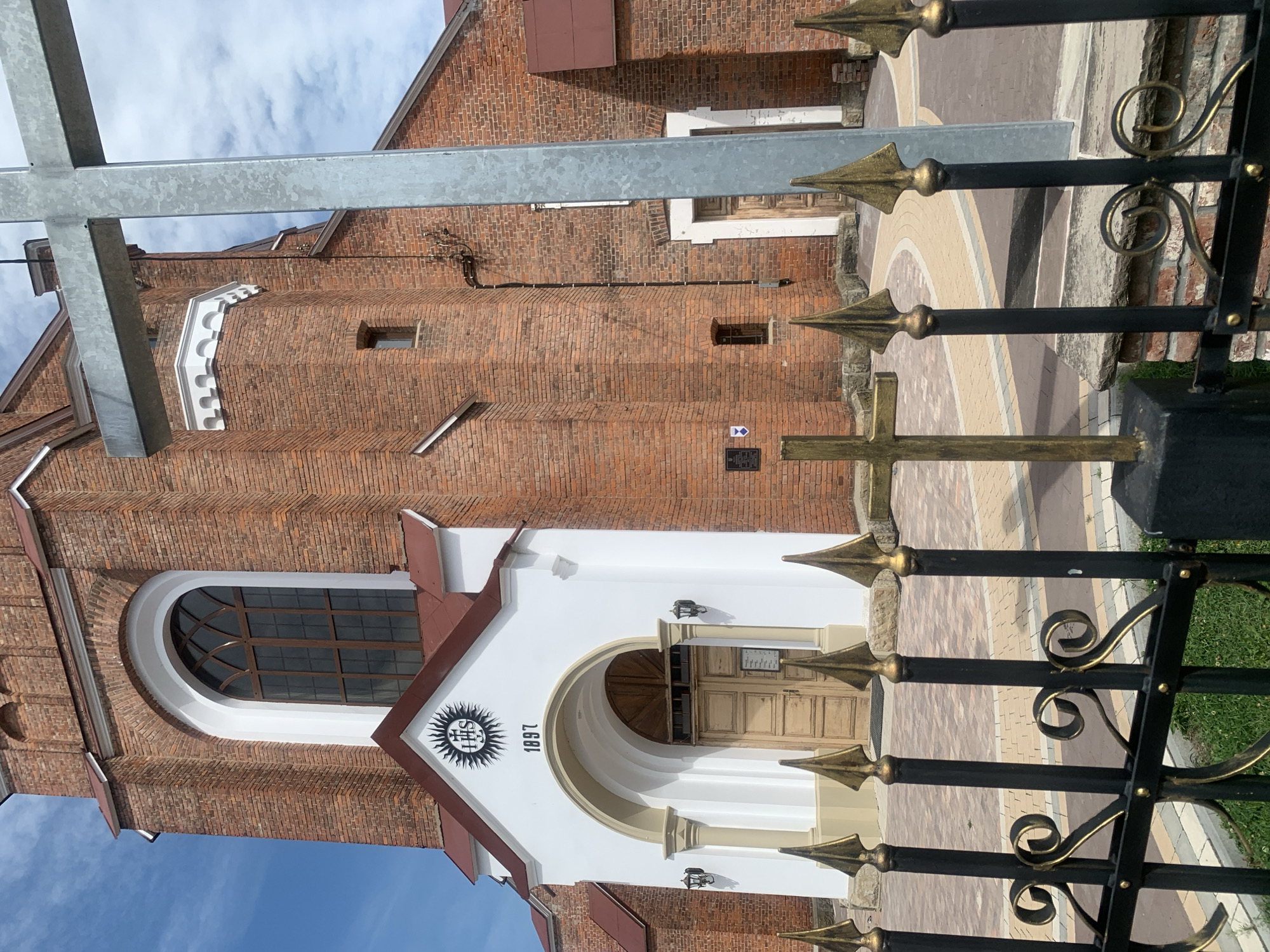The image shows the entrance to a brick church in Kolomyia, Ukraine, on a partly cloudy day. A black metal fence with gold-colored accents runs across the foreground, with a gold-colored cross placed in front of it. Behind the fence, the church has a white facade above the entrance, featuring a large arched doorway with wooden doors and the year "1897" inscribed above it. A religious symbol with the letters "IHS" is also visible on the white facade. The church walls are made of red brick, and a large arched window with dark panes is set high on the facade. A large, silver-colored cross is positioned to the right, partially obscuring the view of the church. The ground in front of the church is paved with light-colored bricks.