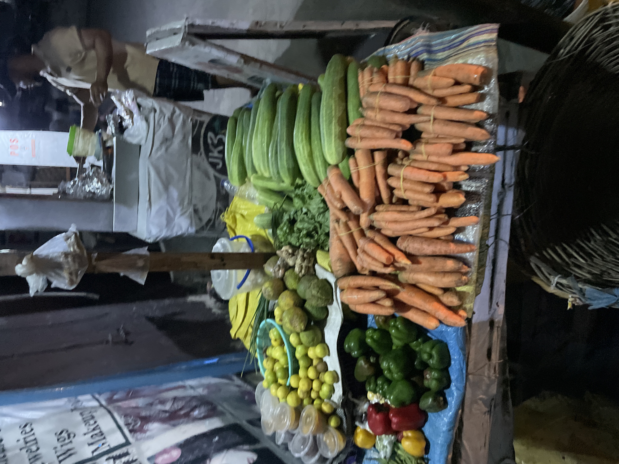An overhead shot captures a bustling market stall in Ado, Nigeria, illuminated at night or late evening. The vendor's display is a vibrant assortment of fresh produce. Dominating the foreground and right are numerous bundles of orange carrots, neatly tied with rubber bands. To their left, a large quantity of long green cucumbers is stacked. Below the cucumbers, a bed of green leafy vegetables, possibly herbs, is visible, alongside fresh ginger roots and a pile of small, round green and yellow fruits, likely limes or similar citrus. Further down, bright green bell peppers are arranged, with a few red and yellow ones adding color to the display.
In the upper left background, a person is partially visible, wearing a light-colored shirt and dark shorts, appearing to attend to the stall or a customer. Various market paraphernalia surrounds the scene, including a white plastic bucket with a blue handle, clear plastic cups, plastic bags, and a large woven basket on the far right. The stall itself appears to be a basic, perhaps temporary, setup.
Visible text includes partial words like "jewelries," "Wigs," and "Makeup" on a white banner in the lower left corner, suggesting other nearby businesses or goods. The letters "JK3" are also visible on a dark object near the center, and "ROSS" on another sign in the background. The overall impression is that of a lively street market operating under artificial light.