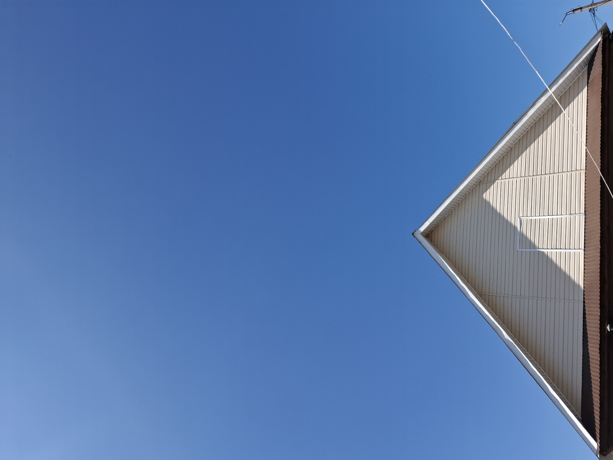 The image shows a building corner against a clear blue sky. The building features light-colored siding, a white trim along the roofline, and a section of dark brown siding on the edge. There appears to be a small, rectangular opening, likely a window, casting a shadow on the siding. A thin wire or cable extends from the top right corner. Based on the information given, the image is taken in Амзя, Russia.