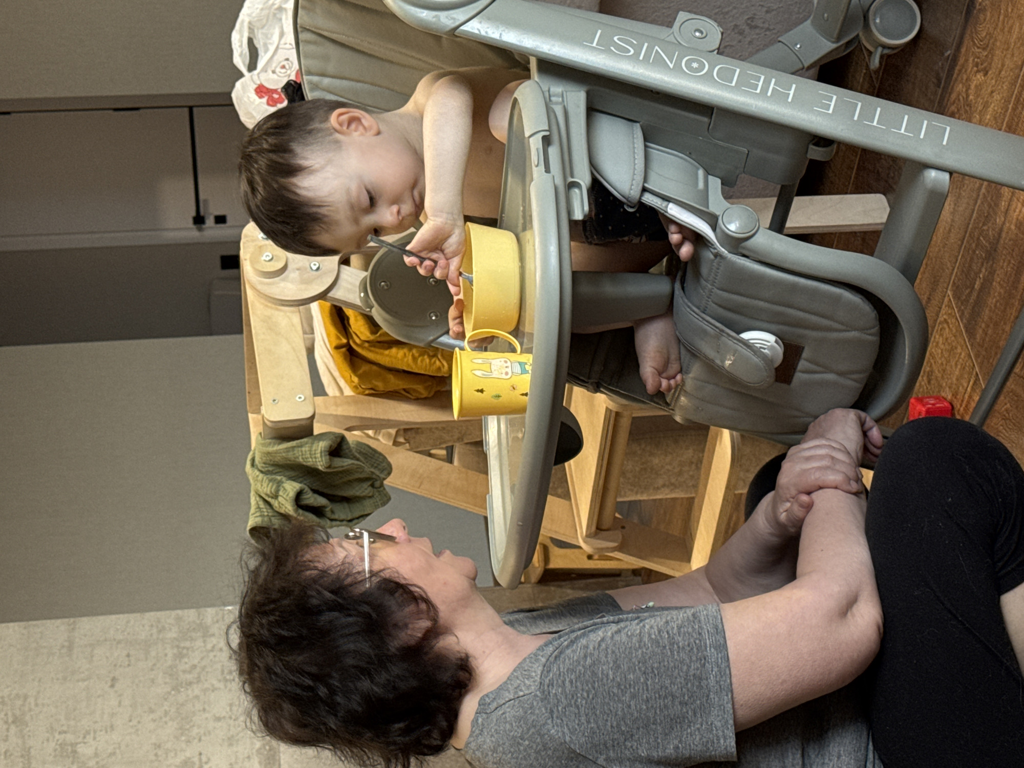 A woman is seated on the floor in an indoor location, interacting with a young child seated in a high chair. The child, with short brown hair, is playing with utensils and cups on the high chair's tray. The high chair is light gray and marked with the words "LITTLE HEDONIST". The woman, with short dark curly hair and glasses, is looking up at the child. She is wearing a gray t-shirt and black leggings. The floor is wood, and the background shows a partial view of a wall and what appears to be a cabinet. There are no indications that this scene is set in Izhevsk, Russia.