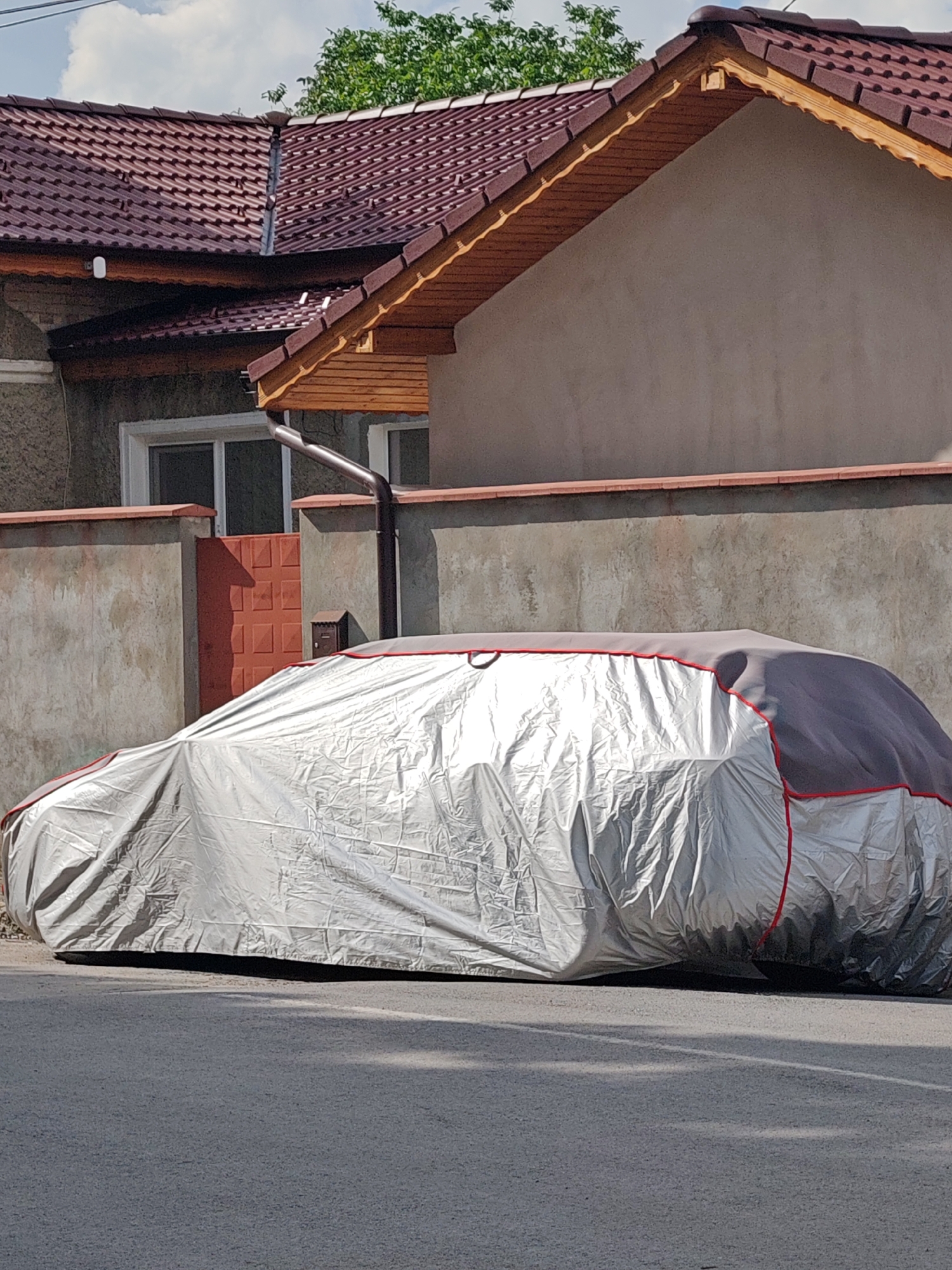 The image depicts an outdoor scene in Tsarev Brod, Bulgaria, under bright daylight. A vehicle, completely covered by a two-tone protective cover, is parked on a dark gray asphalt road in the midground. The car cover is primarily silver with a wrinkled texture, featuring a dark gray upper section and thin red piping along its seams and edges; a single dark loop is visible on the top front of the cover.
Behind the covered car, a low, light gray concrete or stucco wall topped with terracotta-colored coping separates the street from a property. Integrated into this wall on the left is a reddish-brown metal gate with a grid-like embossed pattern. To the right of the gate, a dark brown metal mailbox is affixed to the wall, and a dark brown downspout runs vertically along the wall, connecting to a gutter above. A distinct, elongated human-like shadow is cast onto the wall and gate from the left.
In the background, two residential structures are visible. The prominent house on the right features a light brown stucco facade and a roof constructed from dark reddish-brown corrugated metal tiles. Its eaves are finished with exposed light brown wood paneling and decorative trim. A second, partially obscured structure to the left also has a similar dark reddish-brown roof and a white-framed window set into a light-colored wall. Green tree foliage rises above the rooftops, and the sky is partly cloudy with white clouds against a blue background. No individuals are visible in the scene.