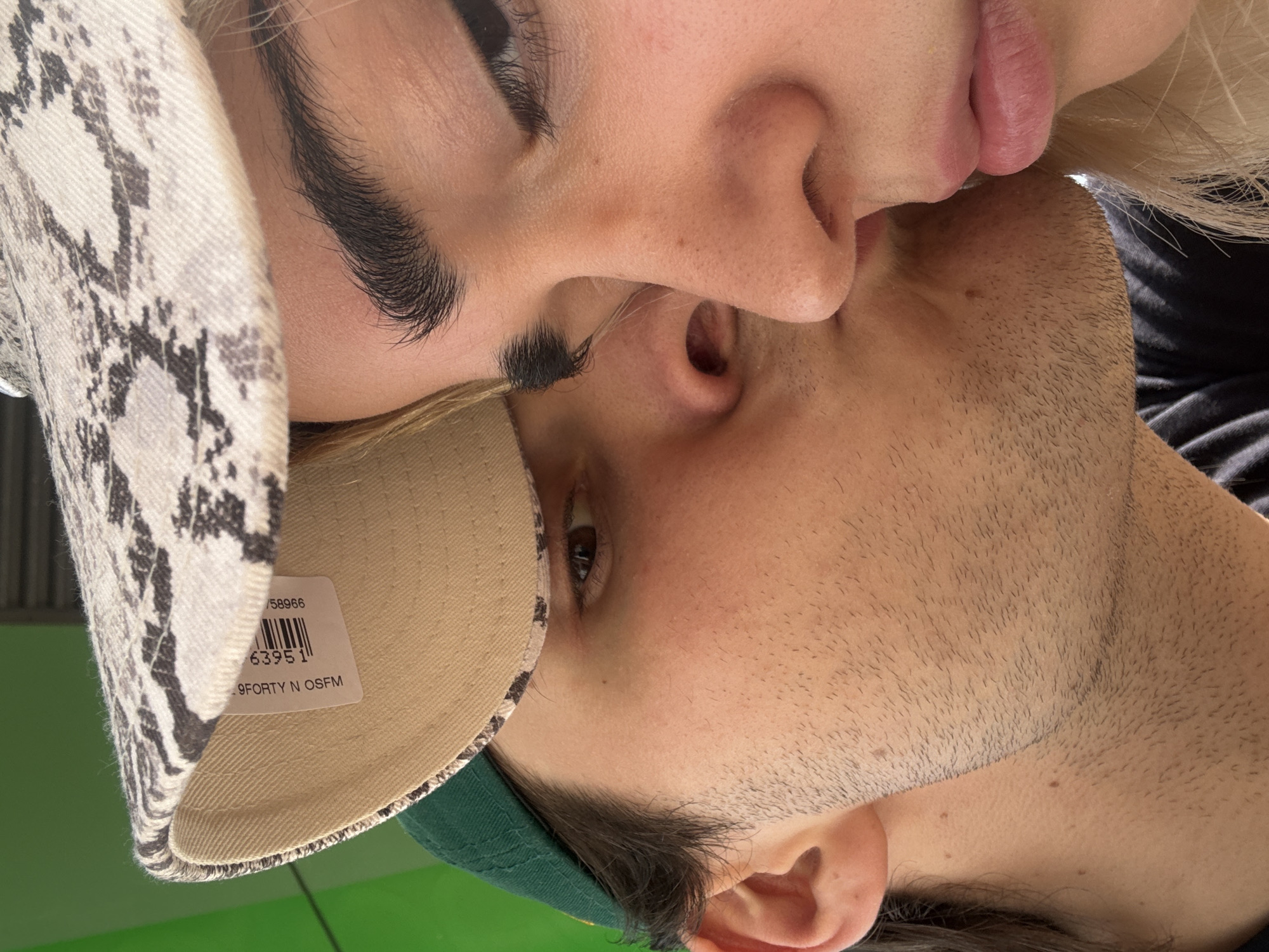 A close-up image depicts a man and a woman in an intimate embrace, likely kissing. The man's face is in the foreground, with a baseball cap featuring a white and grey snake print design visible on his head. The woman's face is partially visible, with her lips pressing against the man's face. The setting appears to be indoors, with a blurry green background. There is no discernible evidence indicating that this image was taken in Milan, Italy.