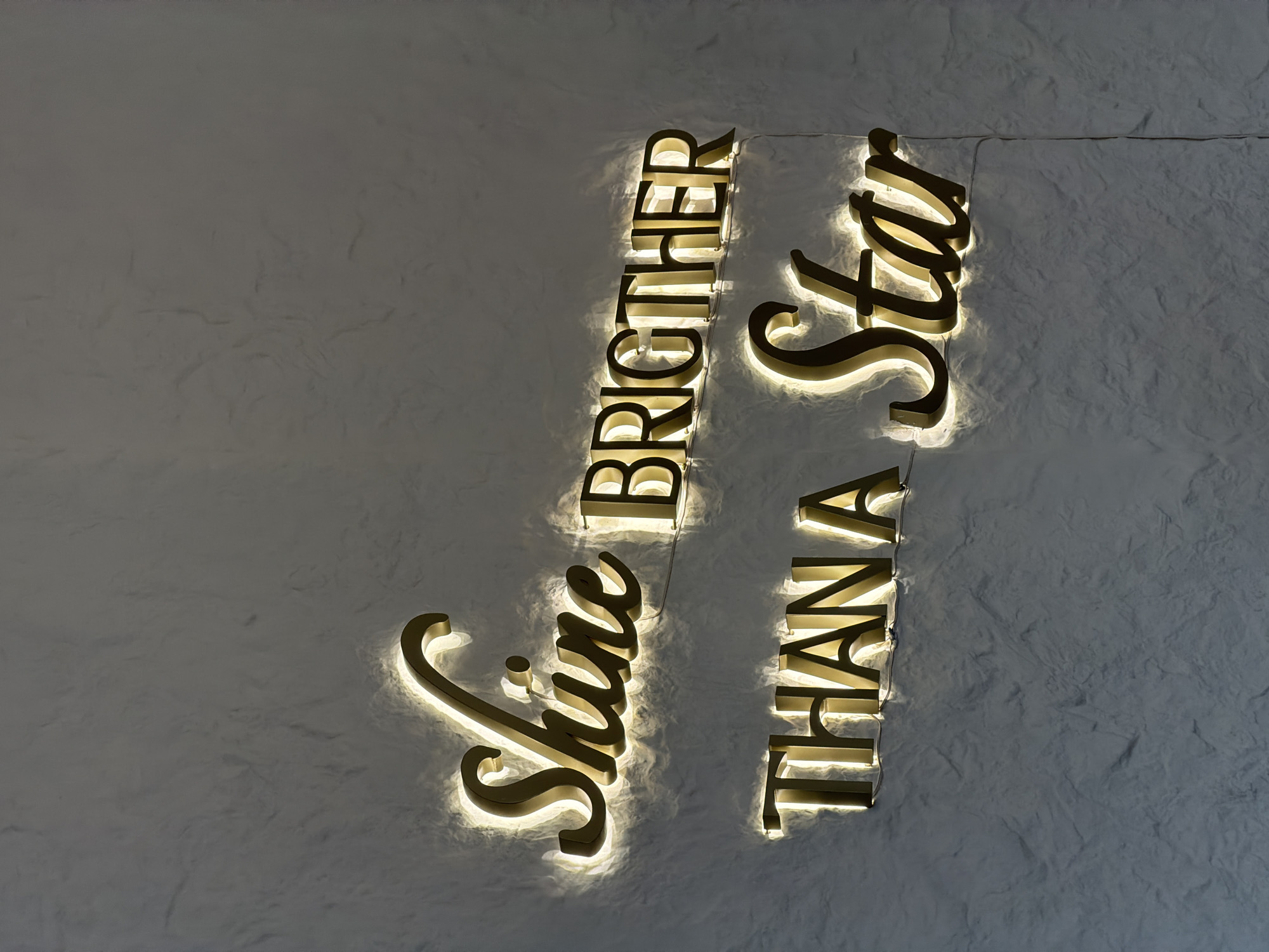 The image displays the words "Shine Brighter Than A Star" mounted on a textured, off-white wall. The letters are gold or brass-colored and appear to be illuminated from behind, creating a glowing effect. Thin wires are visible, connecting the letters and likely providing the power for the lighting. The background wall has a subtle mottled or marbled appearance. The image was taken in North Kuta, Indonesia.