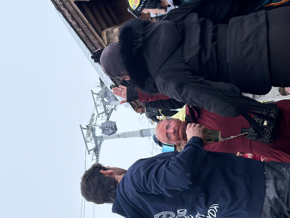 The image captures an outdoor gathering of several people in a cold, possibly snowy environment, characterized by an overcast sky and the presence of a ski lift structure.
In the immediate foreground, with his back mostly to the camera, is a man with dark, curly hair, wearing a dark blue hooded sweatshirt. Part of a white logo and text is visible on the back of his sweatshirt, reading "...L & SKI DAY" along with a circular emblem. His head is turned slightly to his right, and he appears to be looking towards the left side of the frame.
To his right and slightly behind him stands a man facing the viewer, featuring a bald head and light skin. He has a neutral to slight smiling expression. He is dressed in a dark red or maroon jacket or pullover. His right hand is near his chest, holding a small, rectangular object that appears to be a book or case with gold detailing. A black shoulder bag with a textured pattern, possibly with tassels or fringe on the flap, and a gold chain strap, hangs over his left shoulder.
Behind these two figures, a woman is visible, wearing a black winter coat with a prominent black fur-trimmed hood. Her head is covered, perhaps by a dark scarf or part of the hood. She is facing away from the camera, looking towards the left, with her right hand raised as if in a wave or gesture. She is wearing dark gloves. Further behind her and to the right, a person is seen wearing a yellow and red ski helmet with "Red Bull" and "SMITH" logos visible on it.
In the mid-ground and background, a large metal ski lift pylon dominates the left side of the frame, with cables extending across. Several enclosed gondola cabins are suspended from these cables. The sky above is uniformly grey and overcast, suggesting a cloudy day with diffuse lighting. The overall scene implies a ski resort or a mountainous area during winter.