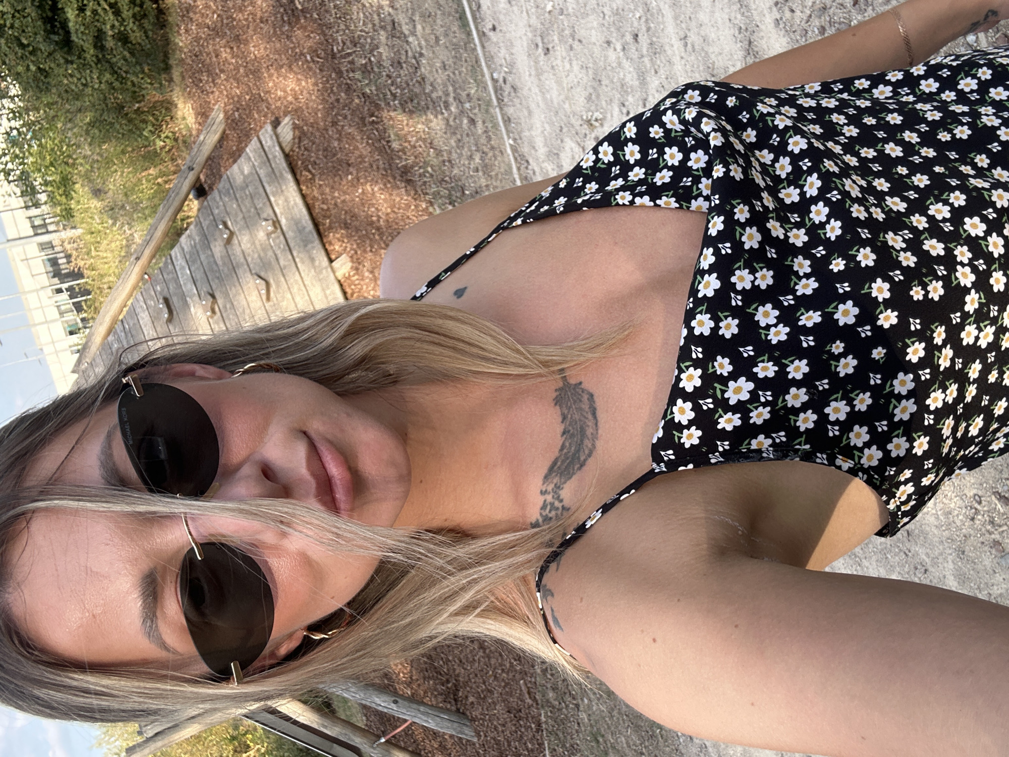 A woman is taking a selfie outdoors. She has blonde hair, is wearing dark sunglasses, and a black dress with a floral pattern. Behind her, a wooden climbing structure is visible. The ground appears to be covered in gravel and dirt. The environment suggests an outdoor recreational area. The woman is positioned in the foreground, with the climbing structure and foliage in the background.