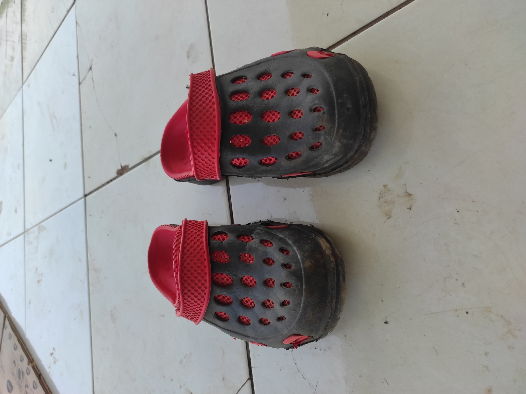 The image shows a pair of black and red clogs placed on a tiled floor. The clogs appear worn and have a perforated design on the upper part, with red material visible underneath the perforations. The tiled floor is light-colored and shows some signs of dirt and minor cracking. In the upper left corner of the image, a portion of a cardboard box is visible. The scene suggests an outdoor or semi-outdoor setting, possibly a pathway or patio. The location is identified as Sumedang, Indonesia.