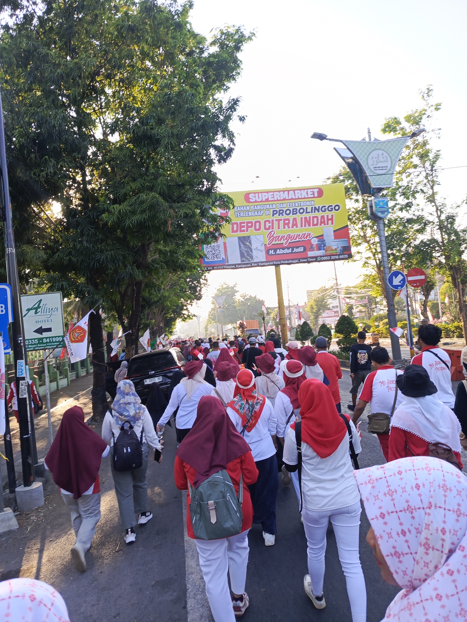 A group of people, predominantly women wearing headscarves and white or red tops, are walking on a paved street, likely during a daytime event. The individuals are mostly facing away from the camera, indicating movement forward along the street. Some are carrying backpacks. A prominent yellow banner hangs overhead, advertising "SUPERMARKET BAHAN BANGUNAN DAN ELEKTRONIK TERLENGKAP DI PROBOLINGGO DEPO CITRA INDAH BANGUNAN H. ABDUL JALIL". This suggests the location is Semampir, Indonesia, near Probolinggo. Several flags with red and white designs are visible, attached to poles along the side of the road. Trees line the street, and a street lamp with an Islamic motif is also present. The sun is shining, casting shadows on the ground. A black car is parked on the right side of the street. The overall scene depicts a procession or organized walk in a semi-urban environment.